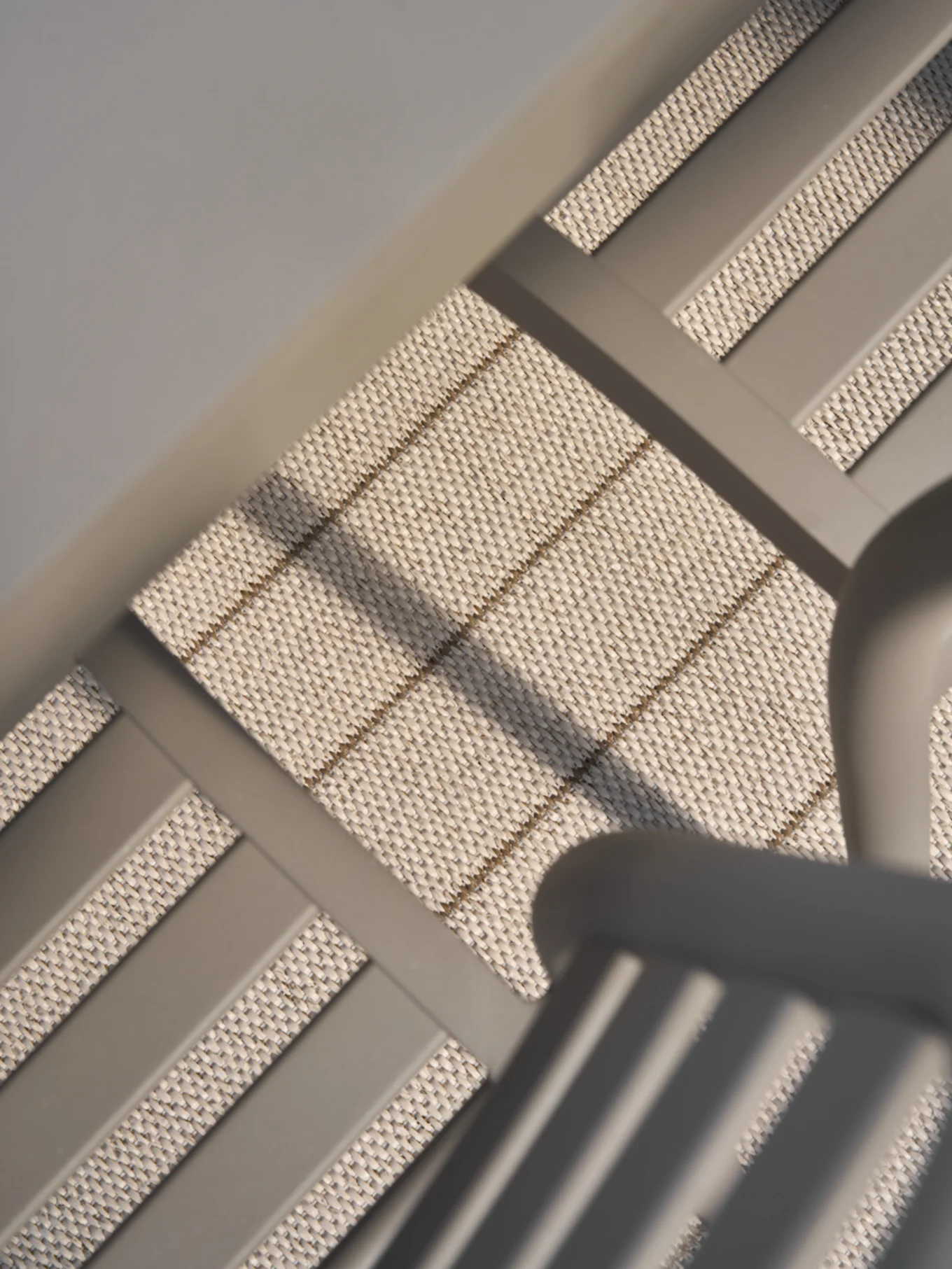 Närbild, vinklad vy av ljust beige vävt material med subtila horisontella och diagonala bruna ränder, inramat av släta grå stänger.