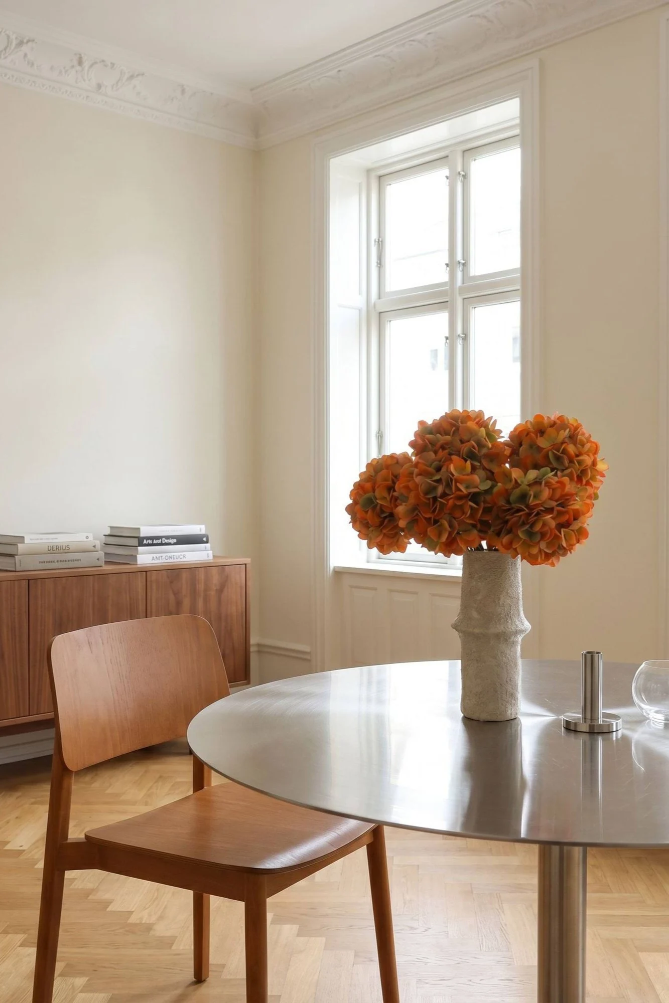 Ett ljust matsalsrum med ett runt metallbord, en trästol och en vas med orange blommor.