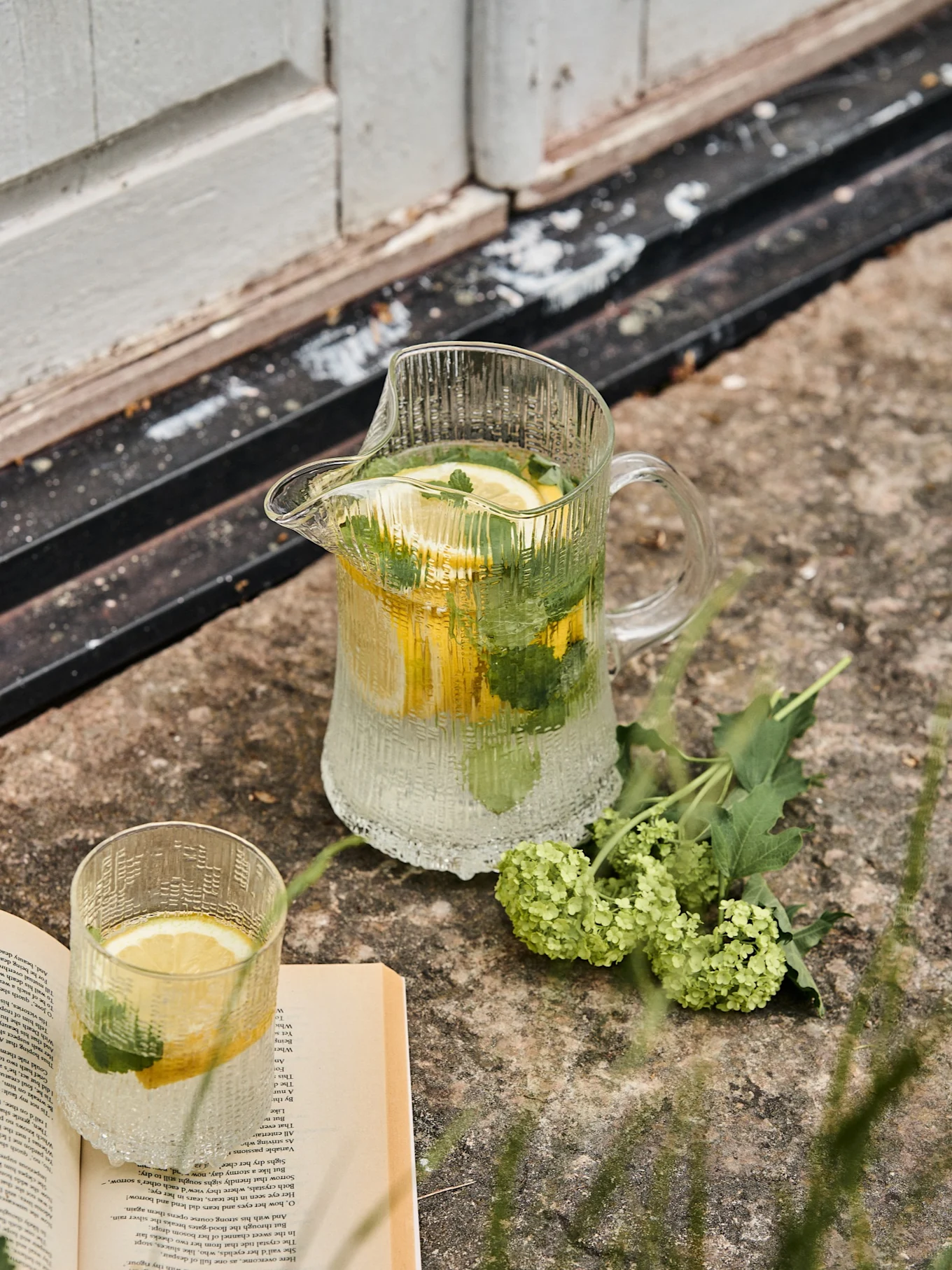 Räfflad glaskanna och matchande glas med citron- och myntavatten, bredvid en öppen bok och gröna blommor på sten.