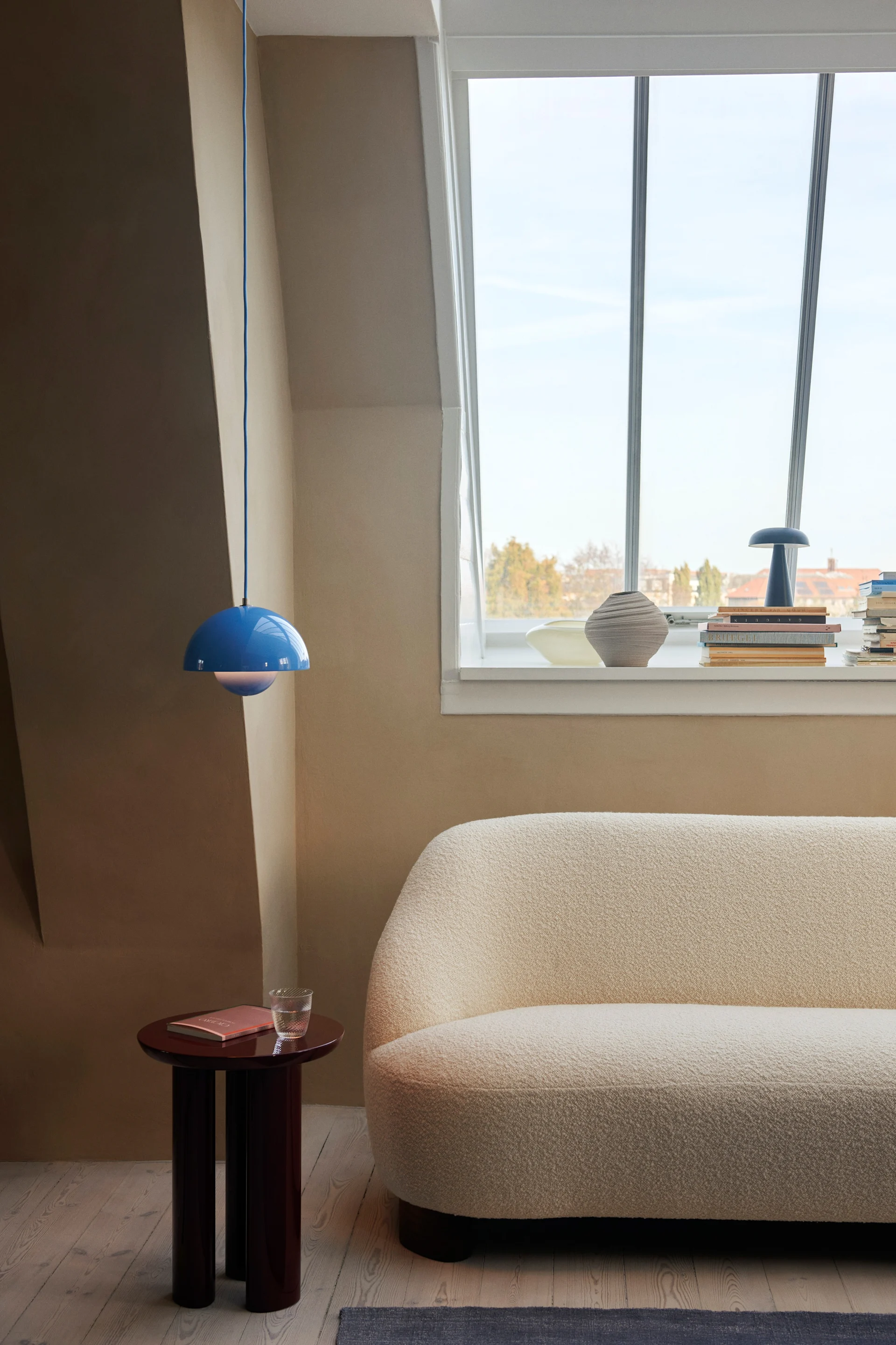 Flowerpot pendellampa VP1 i en klar blå färg hänger i taket i ett naturfärgat rum, på bilden syns även ett sidobord i mörkt trä och en soffa i naturfärg. 