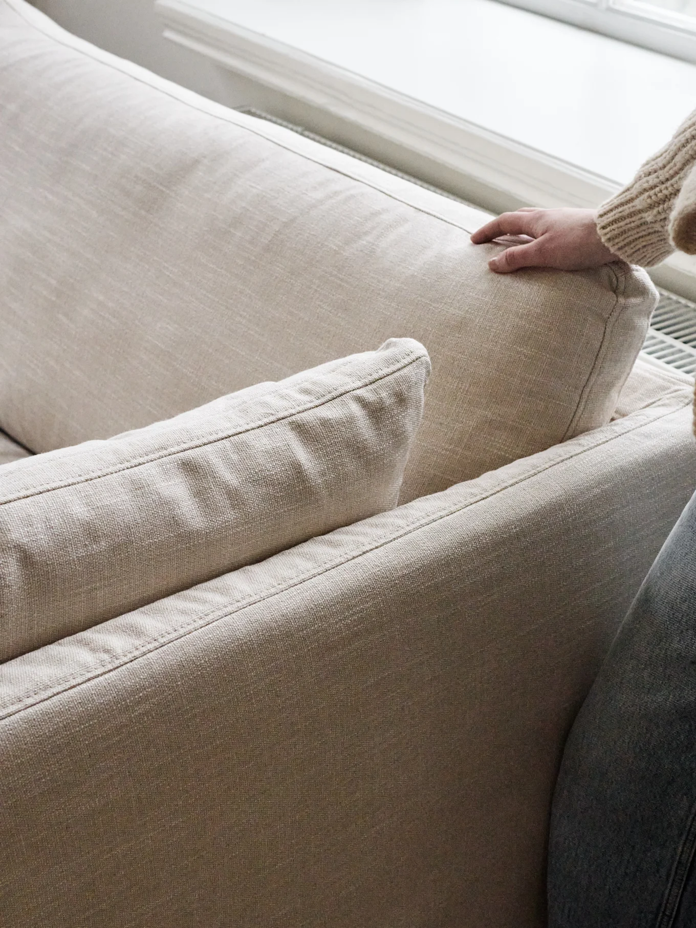 En hand i en grovstickad tröja trycker försiktigt ner i ryggkudden på en ljusbeige soffa.