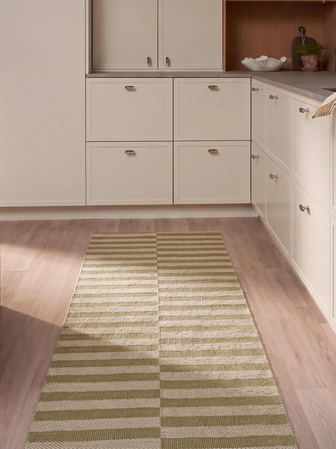 Ett kök med ljusa skåp, trägolv och en lång rektangulär matta med gröna och vita horisontella ränder.