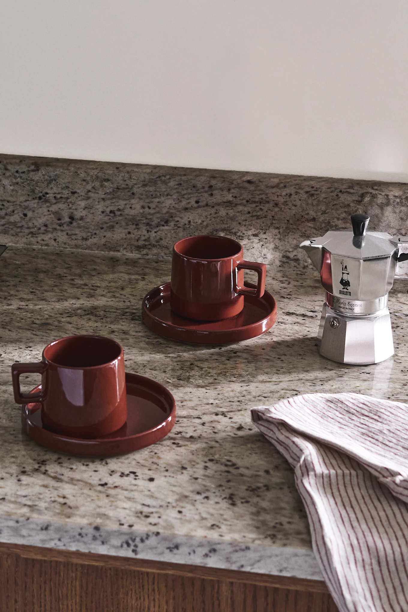 Två röda keramikmuggar och en silverkaffebryggare på en fläckig granitbänkskiva med en randig handduk.