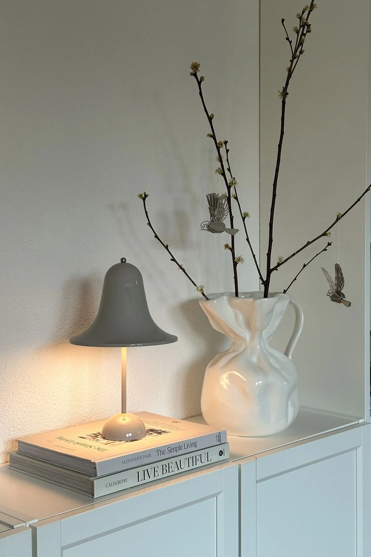 På @oursmall.home kan du se två möjliga Mors dag-presenter: Pantorp bordslampa från Verpan står på en byrå, bredvid den Crumple vas från Byon med några kvistar i.