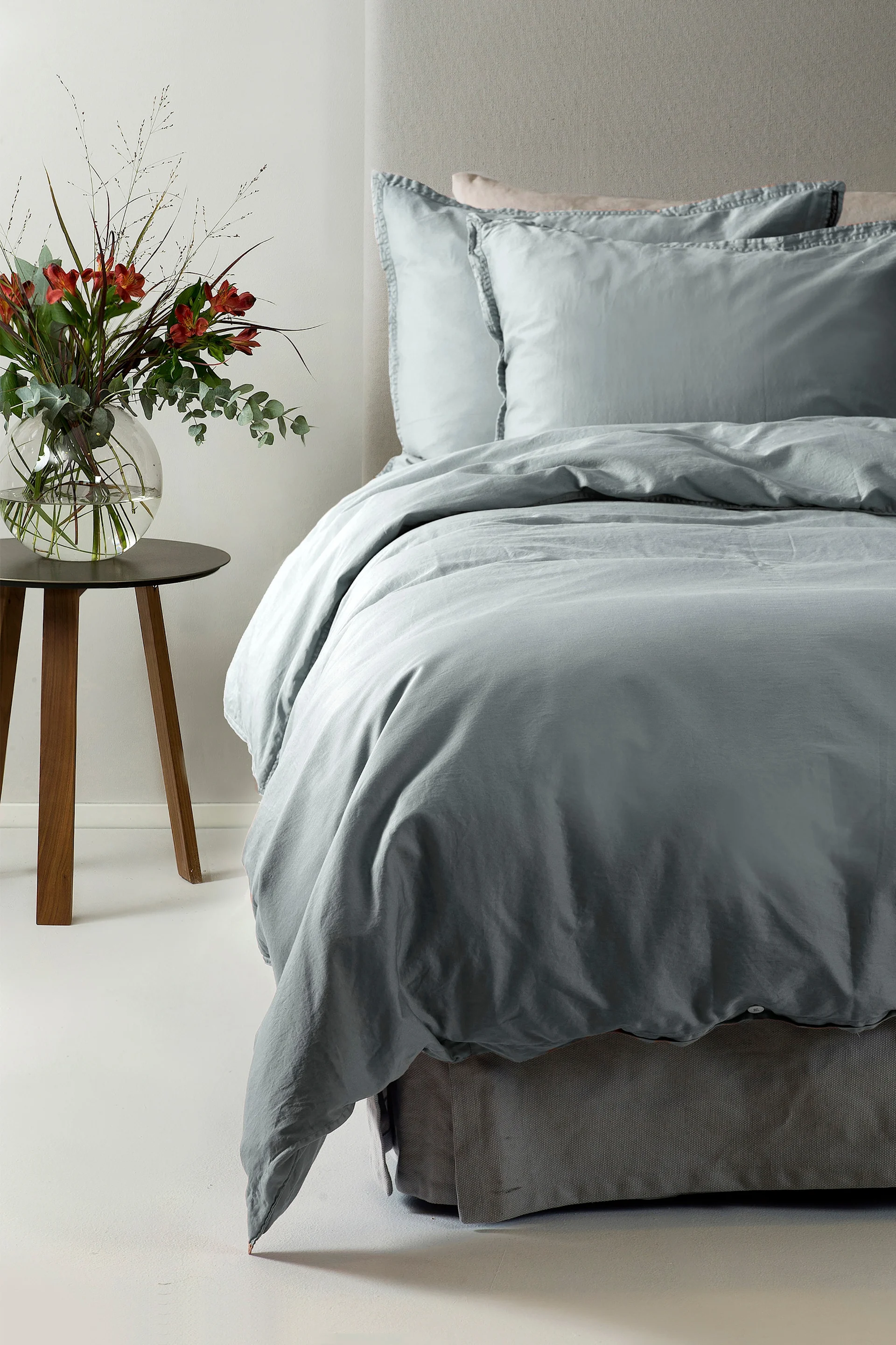 En säng med ljusblå sängkläder och två kuddar, bredvid ett träbord med en vas med röda blommor.