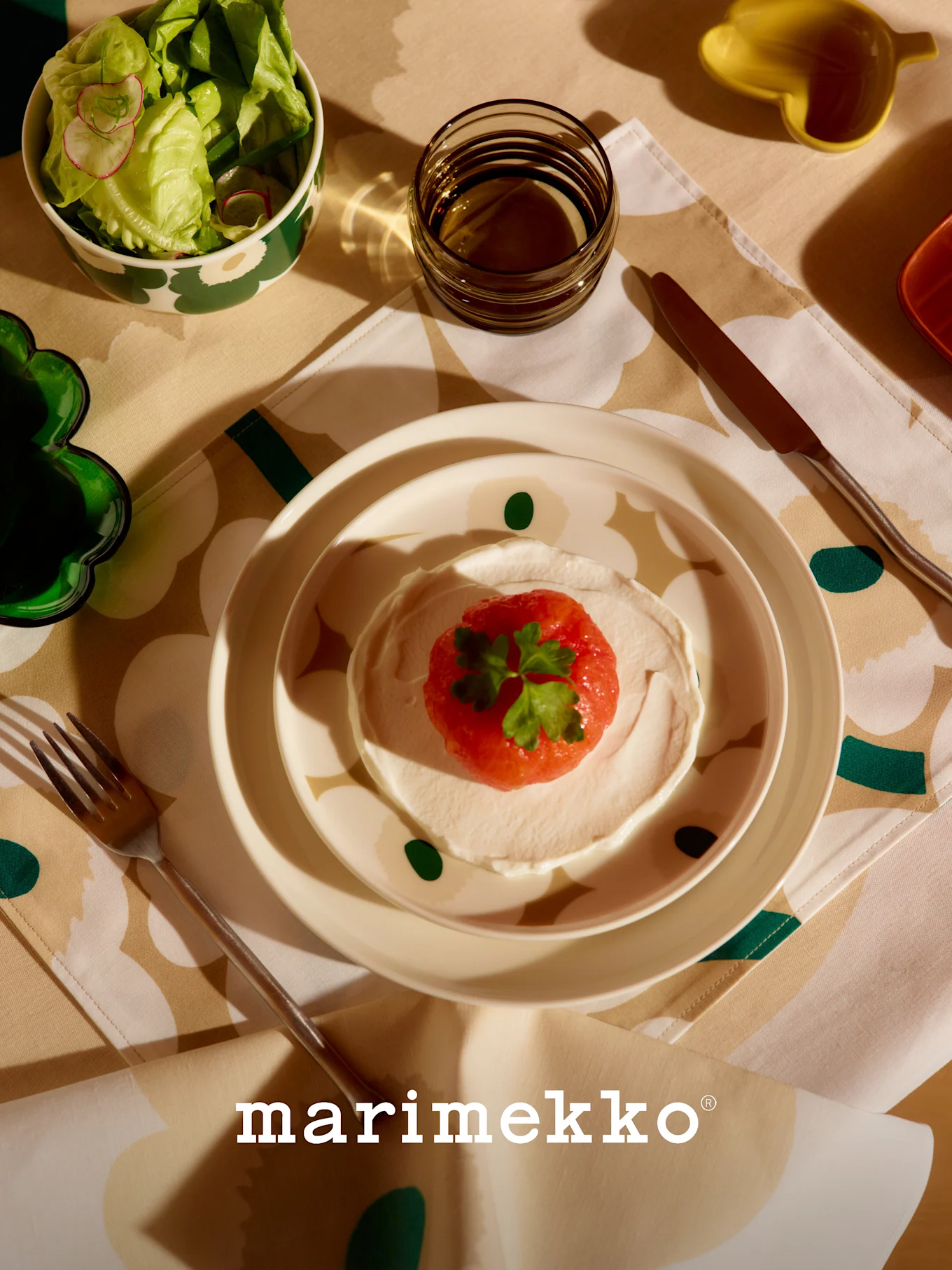 Marimekko borddukning med mönstrad salladsskål, rätt med röd frukt och grädde, glas och bestick på en blommig duk.