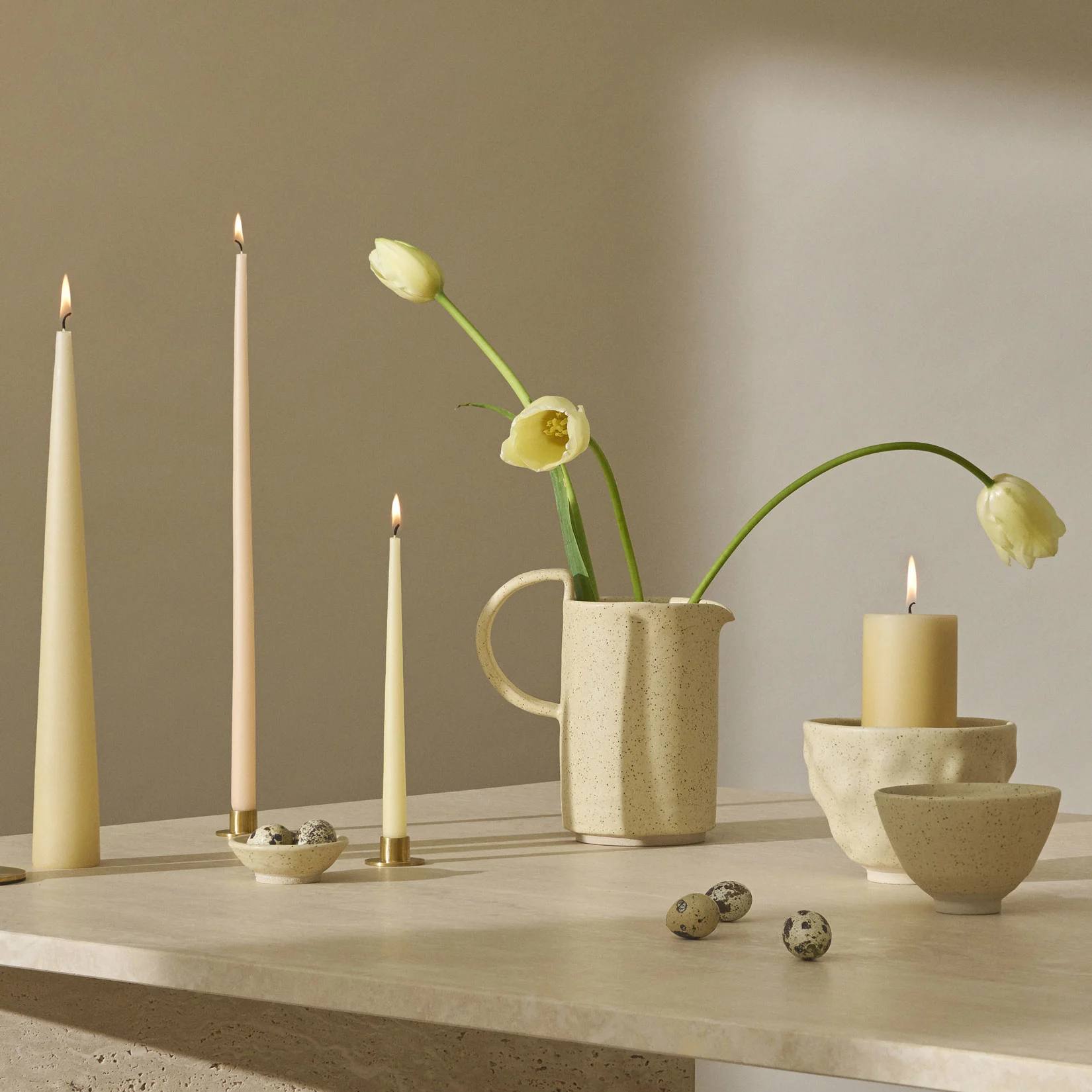 Ett stilleben med tända ljus, ljust gula tulpaner i en spräcklig kanna och vaktelägg på en texturerad yta.