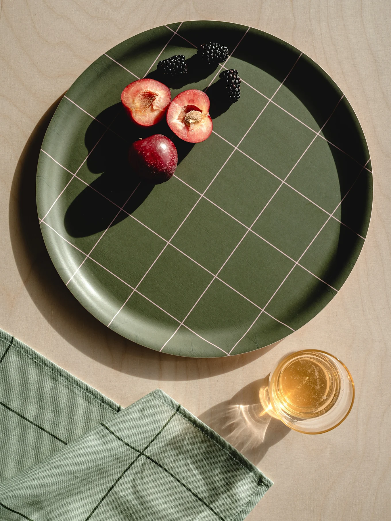 Mörkgrön rutig bricka med röda plommon, björnbär, ljusgrön servett och ett glas juice på trä.