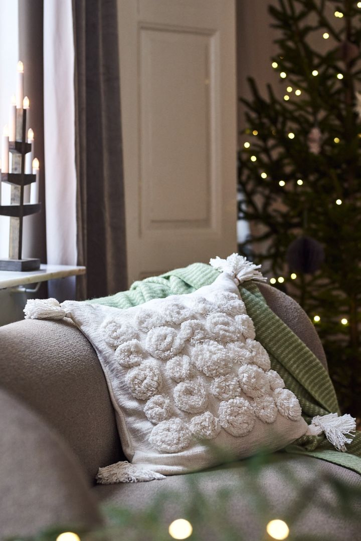 Vit kudde med tofsar och en grön pläd i soffan blir stilsäker och modern julinredning i år. 
