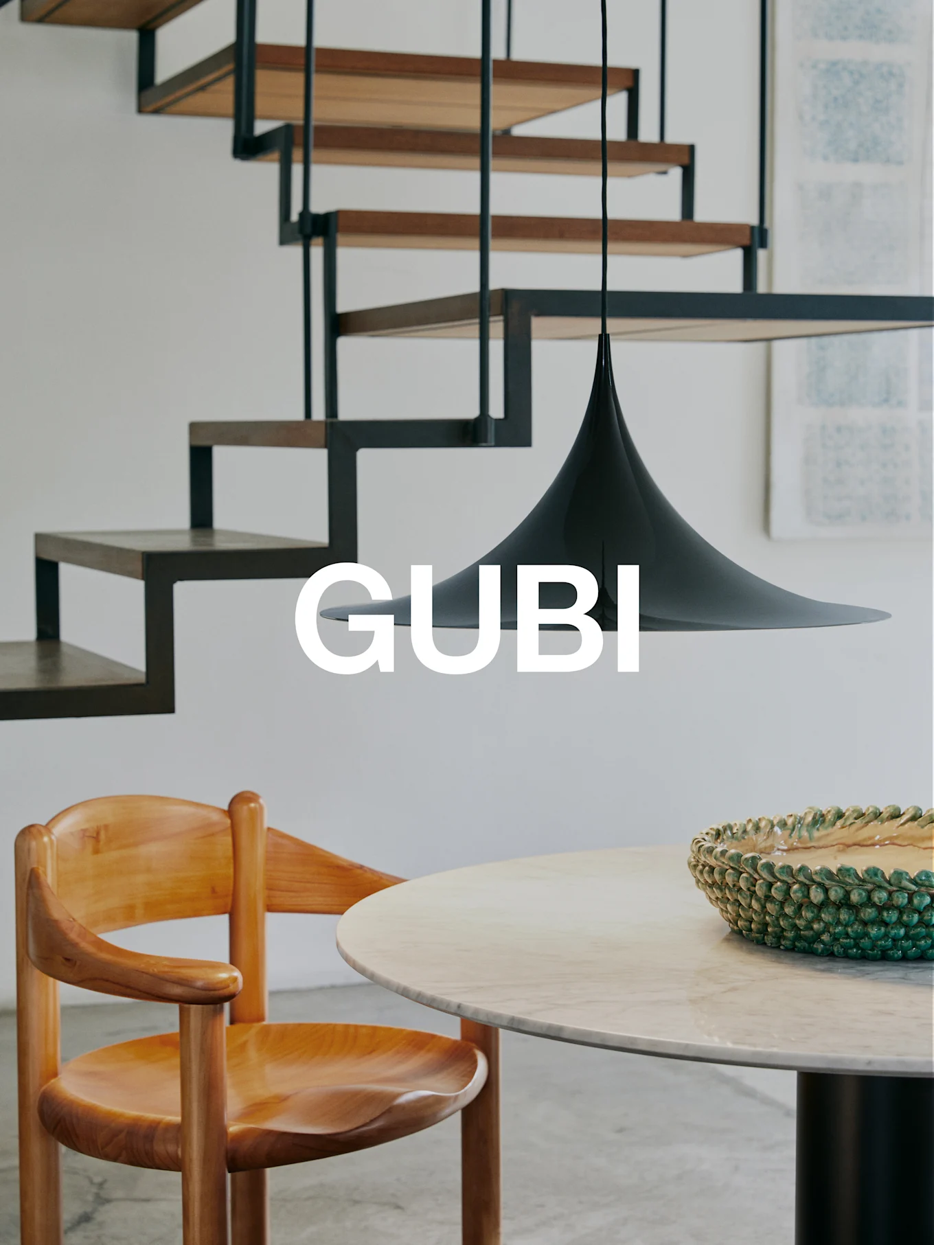 GUBI-logotyp i en modern inredning med trästol, marmorbord, grön skål, svart pendellampa och en modern trappa.