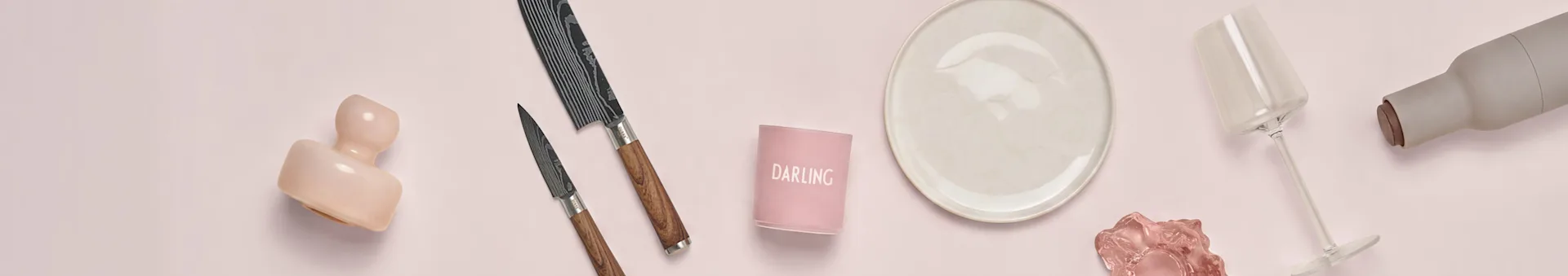 Dekoration på rosa: rosa vas, mönstrade knivar, 'DARLING' ljushållare, beige tallrik, vinglas, grå flaska.