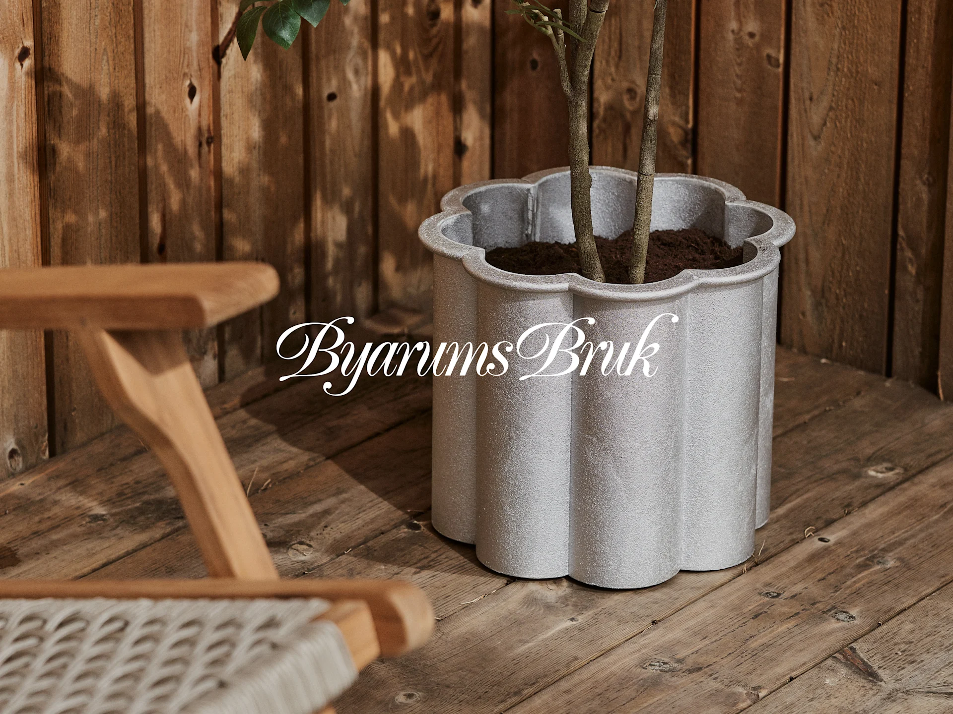 En silvergrå vågig planteringskruka med en växt på ett trädäck, bredvid en trästol. Logotypen 'Byarums Bruk'.