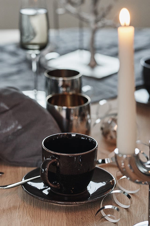 Rustik, svart kaffekopp med fat från Broste Copenhagen skapar kontrast på en nyårsdukning i silver. 