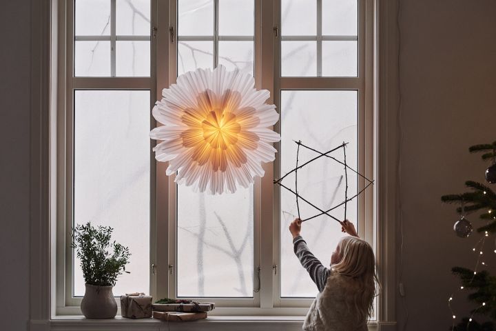 Snöblomma adventsstjärna från Watt & Veke i ett fönster, barn bredvid som håller i en stjärna av pinnar. 