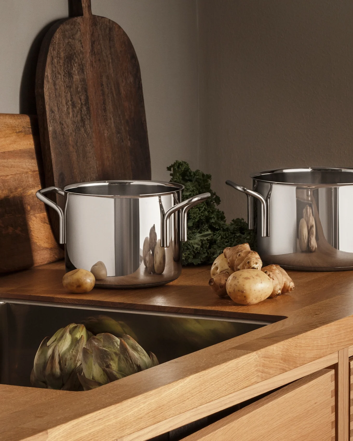 Modernt kök med träbänkskiva, diskho i rostfritt stål, kran, tre kastruller och färska råvaror som potatis och kronärtskockor.