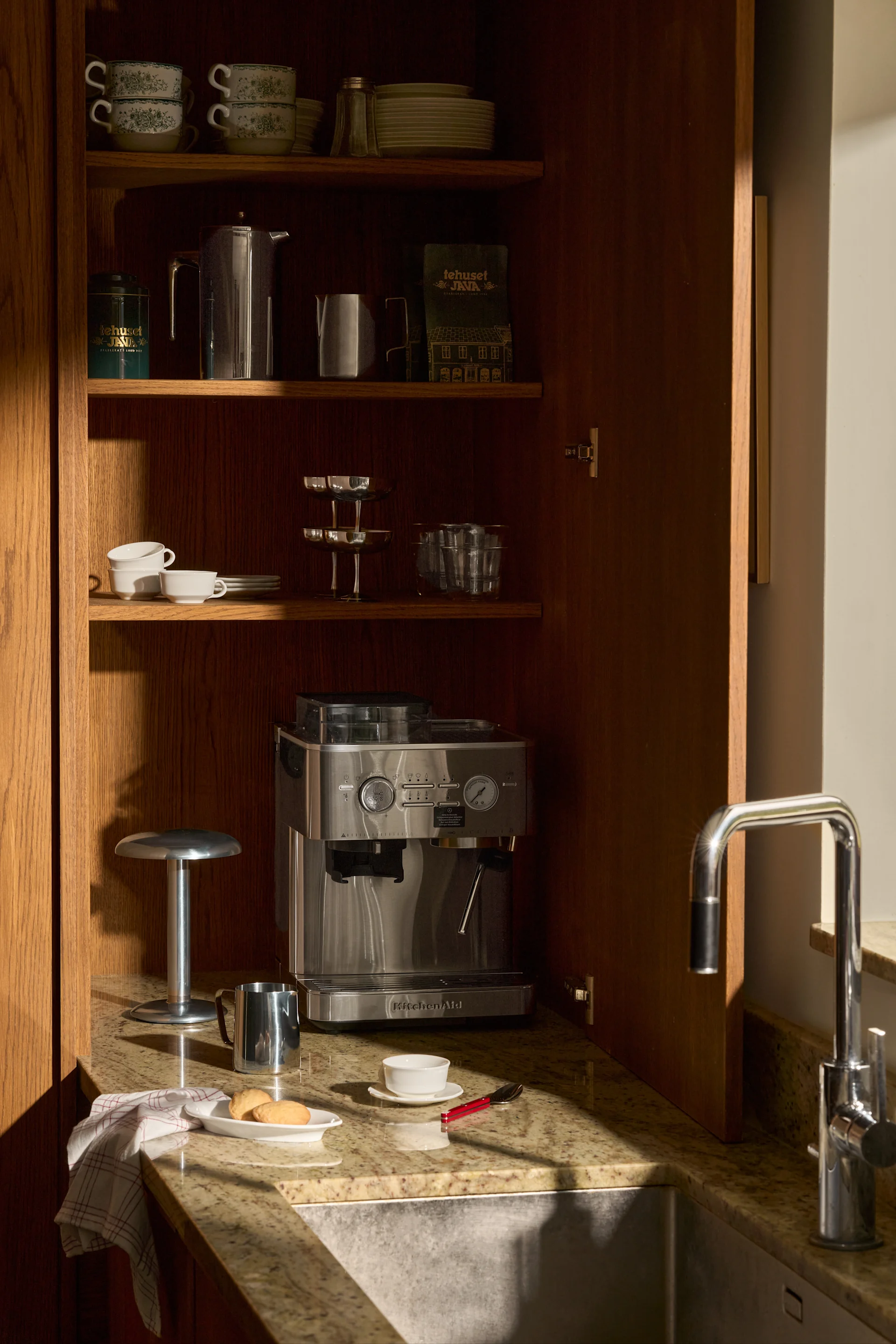Öppet träskåp: KitchenAid espressomaskin, staplade koppar, tallrikar, kaffe och diskho på granitbänkskiva.