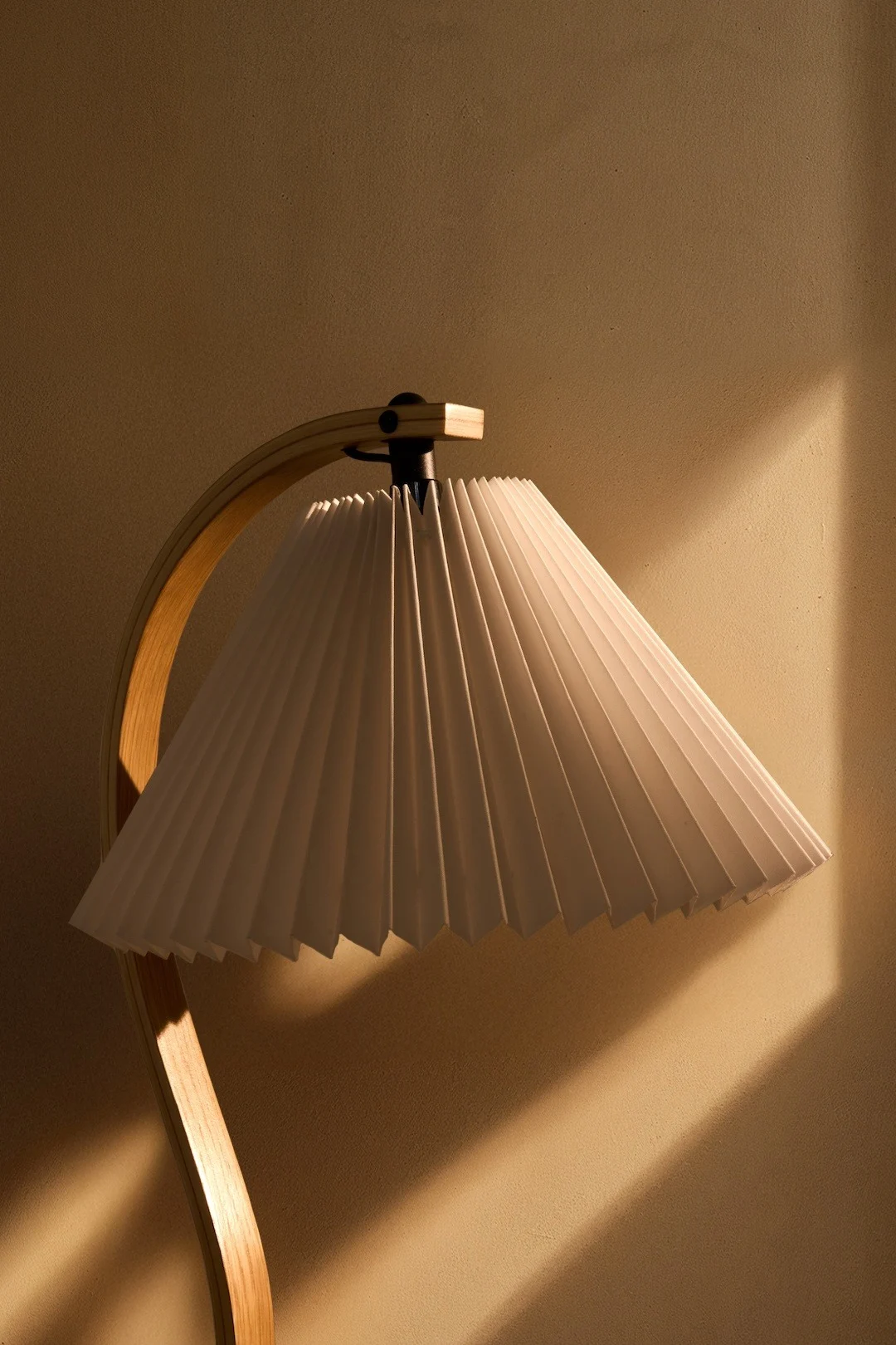 En lampa med en vit plisserad skärm och en böjd träfot, som kastar varmt ljus på en texturerad vägg.