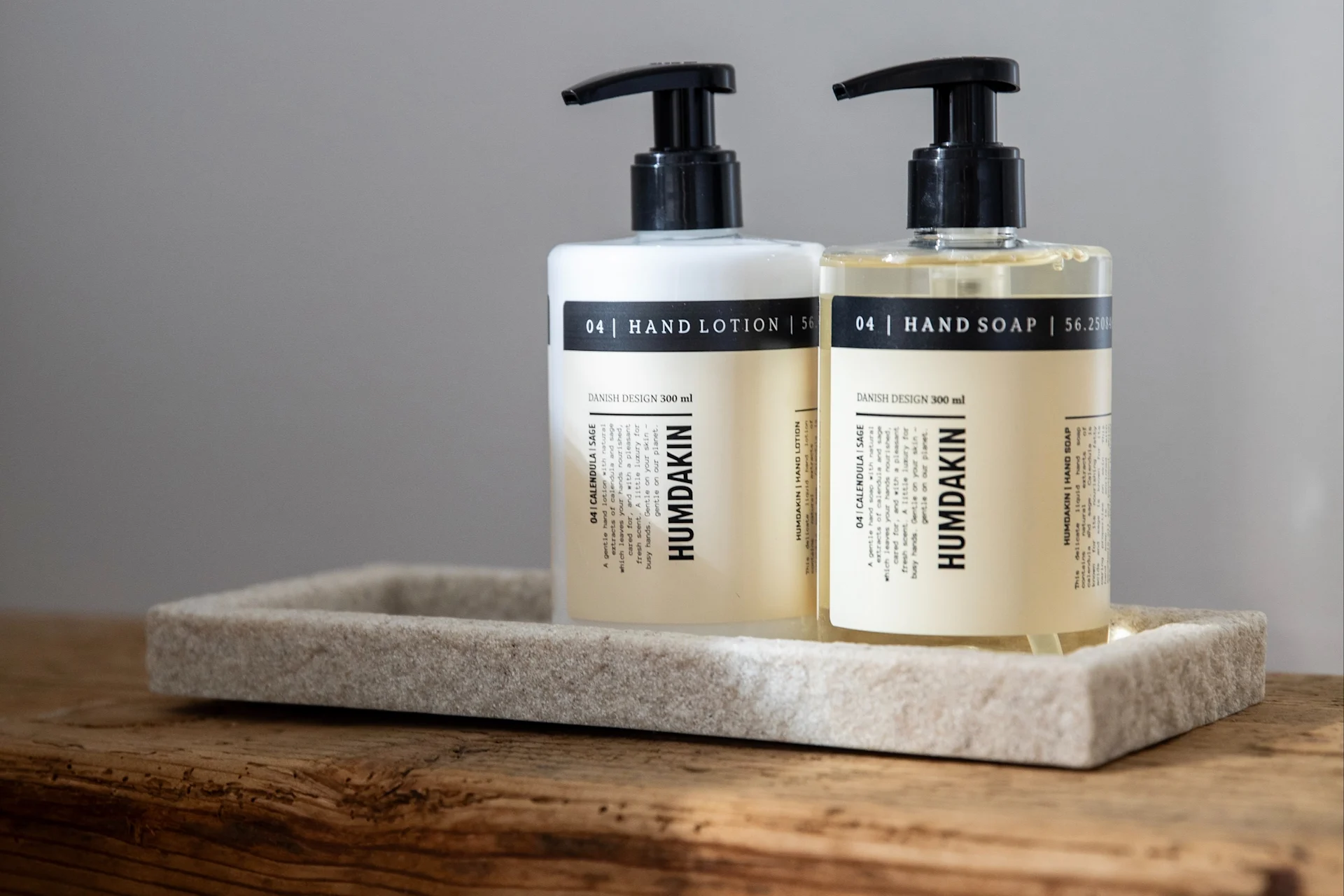 Två Humdakin pumpflaskor, handlotion (vit) och handtvål (klar), på en stenliknande bricka på trä.