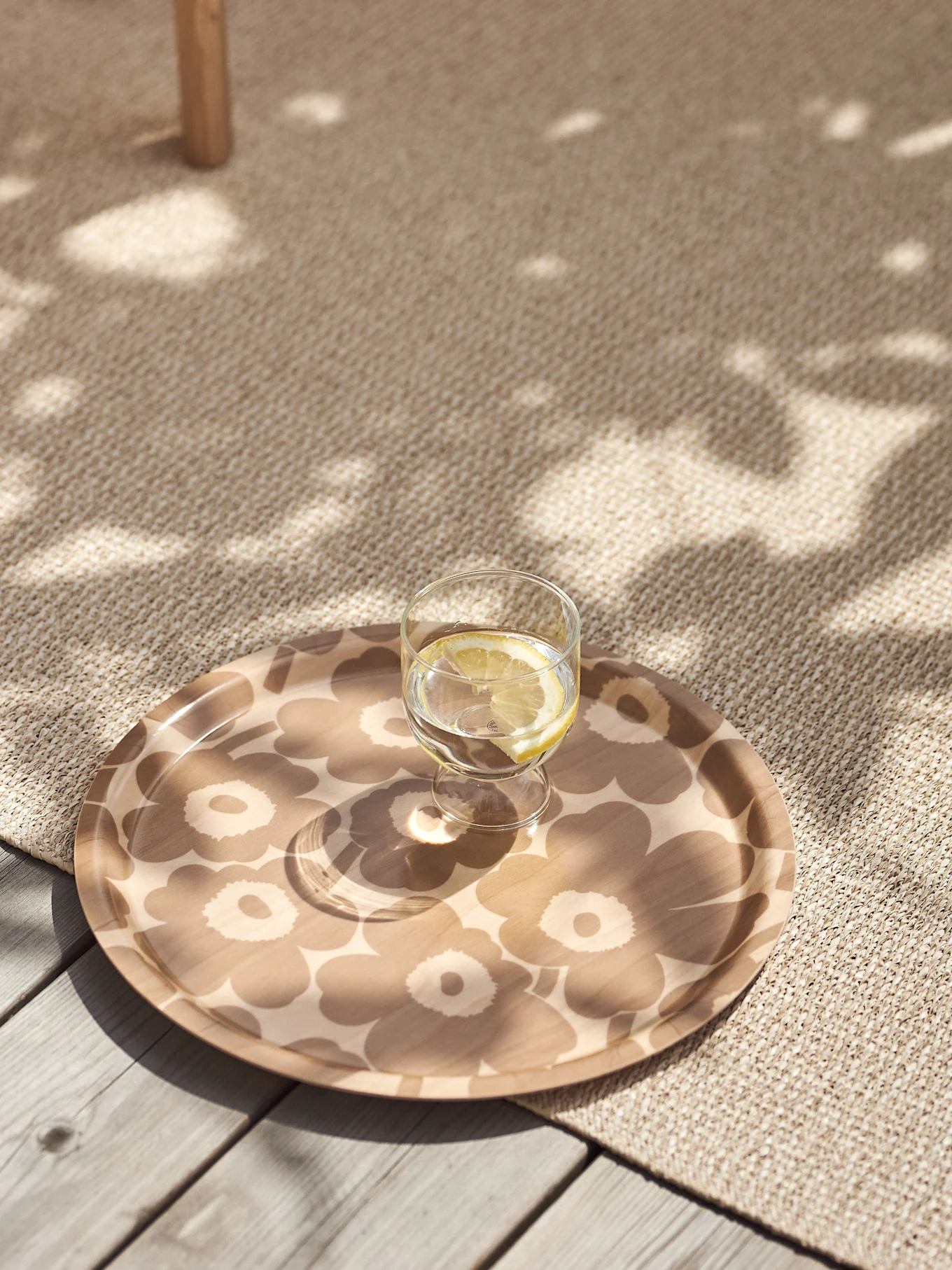 Glas med citronvatten på en beige blommönstrad bricka på en texturerad matta med fläckigt solljus.