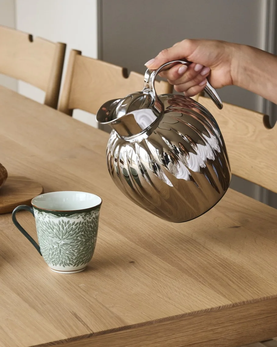 En hand häller från en silverfärgad räfflad karaff i en vit mugg med grönt bladmönster på ett träbord.