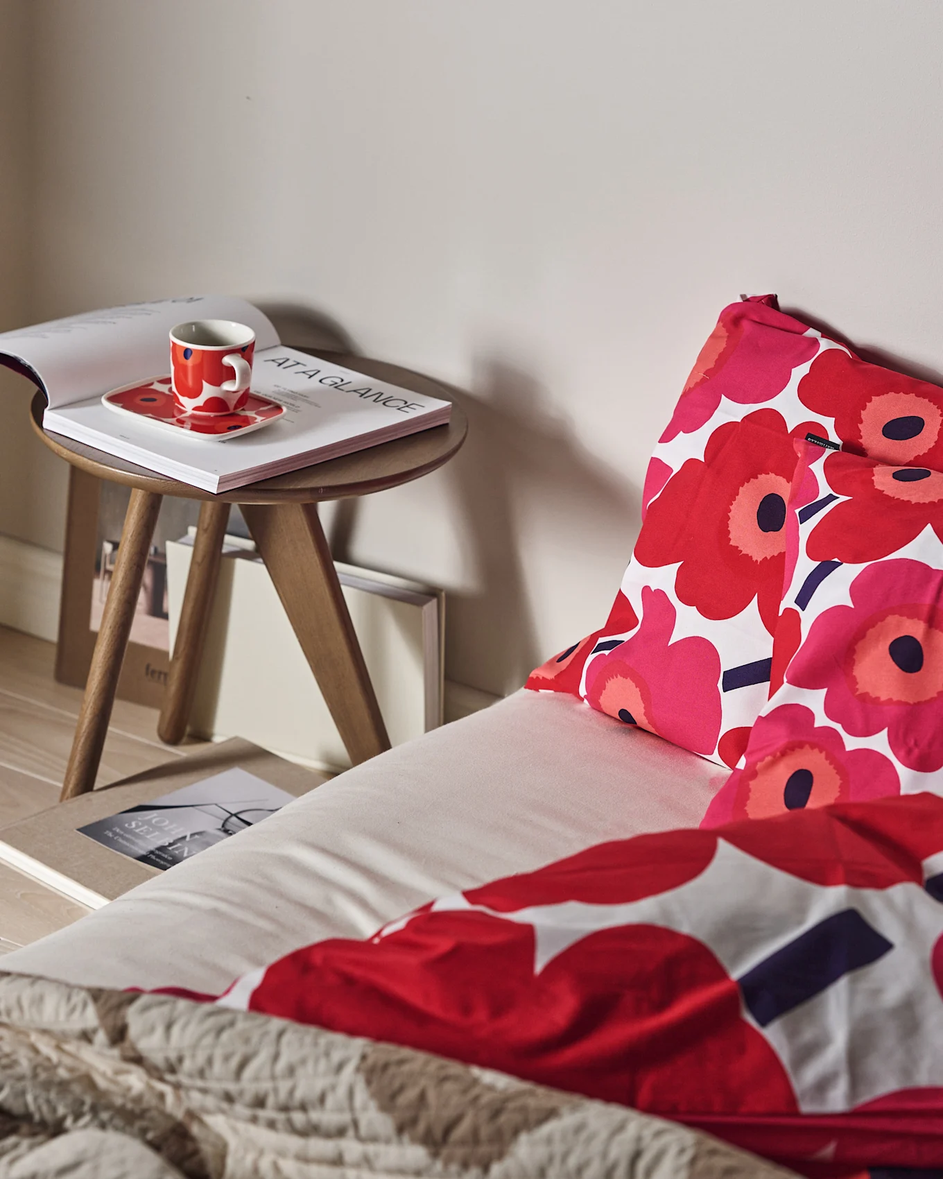 En säng med rött och rosa blommigt sänglinne och en texturerad beige pläd. En röd kopp och öppen bok på ett sidobord i trä.