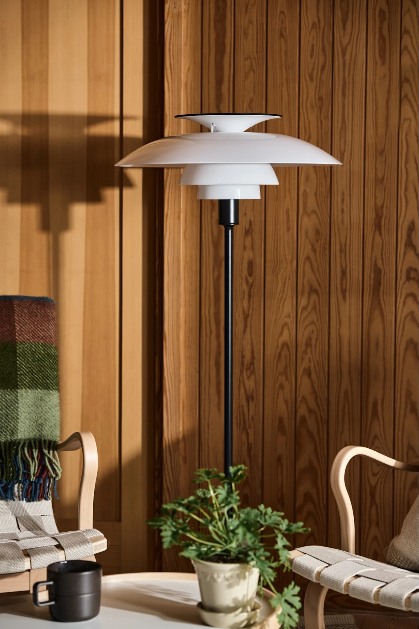 Vit skiktad golvlampa bredvid två trästolar och en växt mot en träpanelvägg.