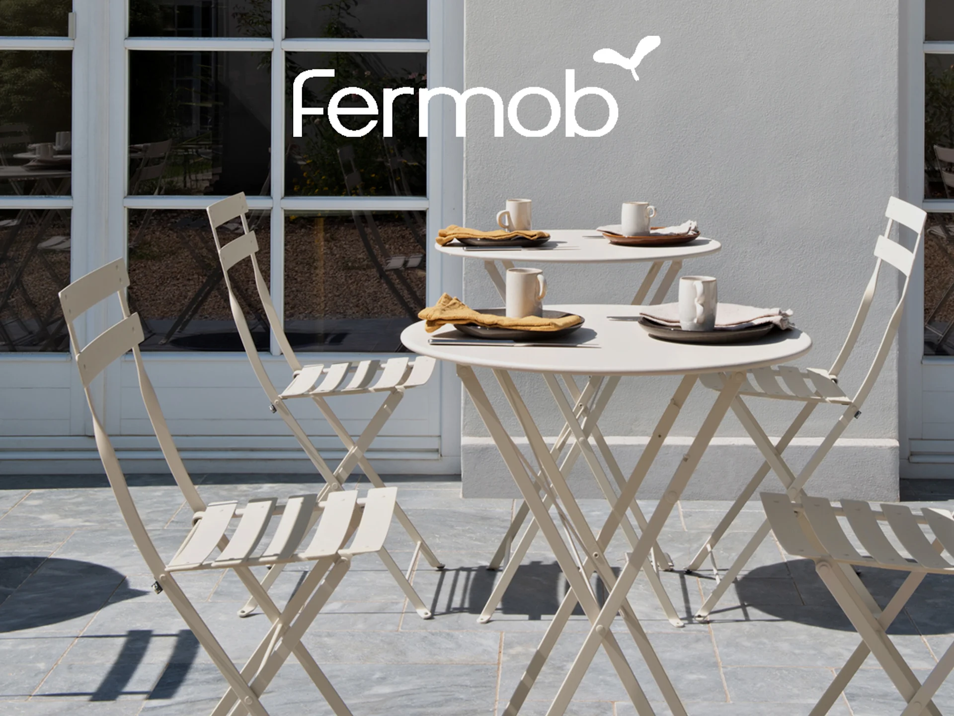 Fermob-logotyp över två utomhuskaffebord, vardera med muggar, tallrikar, servetter, omgivna av matchande stolar på en uteplats.
