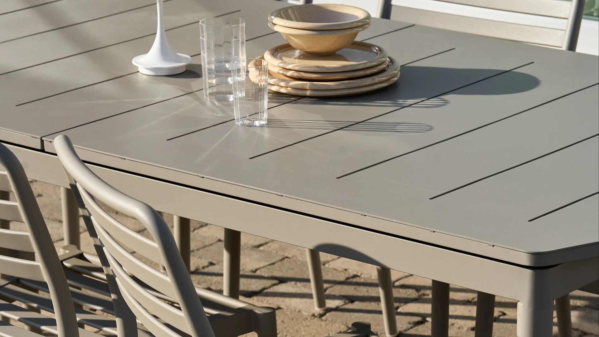 Ljusgrått lamellutemöbel med staplade beiga tallrikar och skålar, två klara glas och en vit prydnadsföremål.