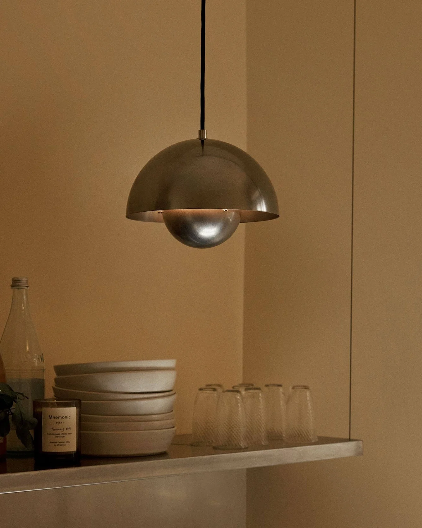 En kupolformad metallpendellampa hänger över en hylla med skålar, glas, flaskor, en frukt och ett doftljus.