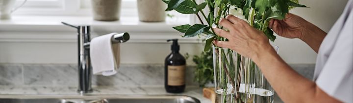 Alvar Aalto-vasen i färgen clear från Iittala står vid en vask i ett kök, innehållandes gröna blomster.