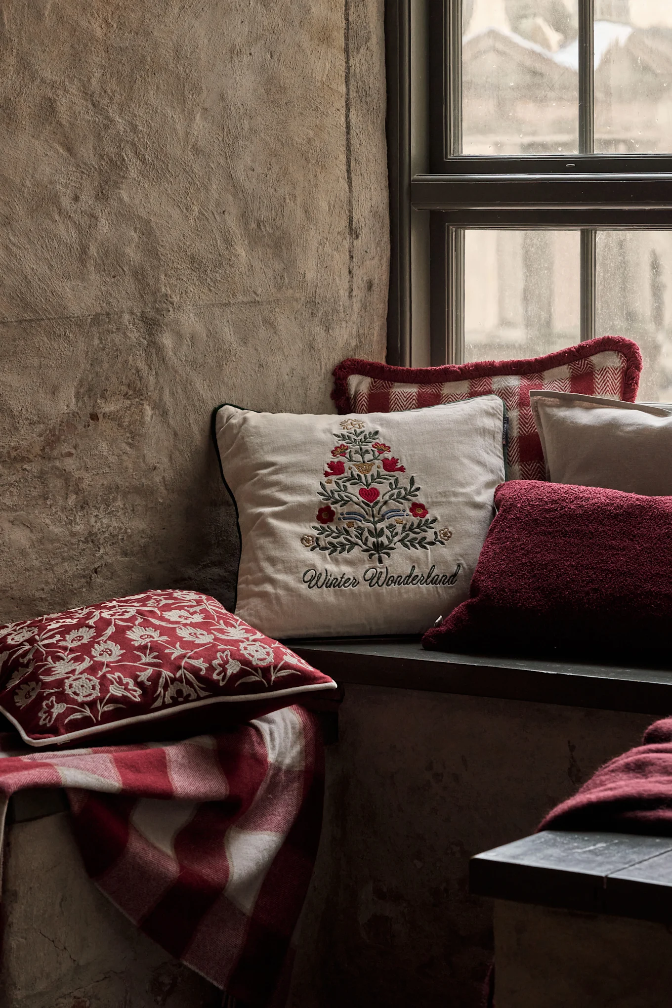Mysig fönsternisch med festliga kuddar, inklusive en med broderad julgran och 'Winter Wonderland', samt en rödrutig filt.