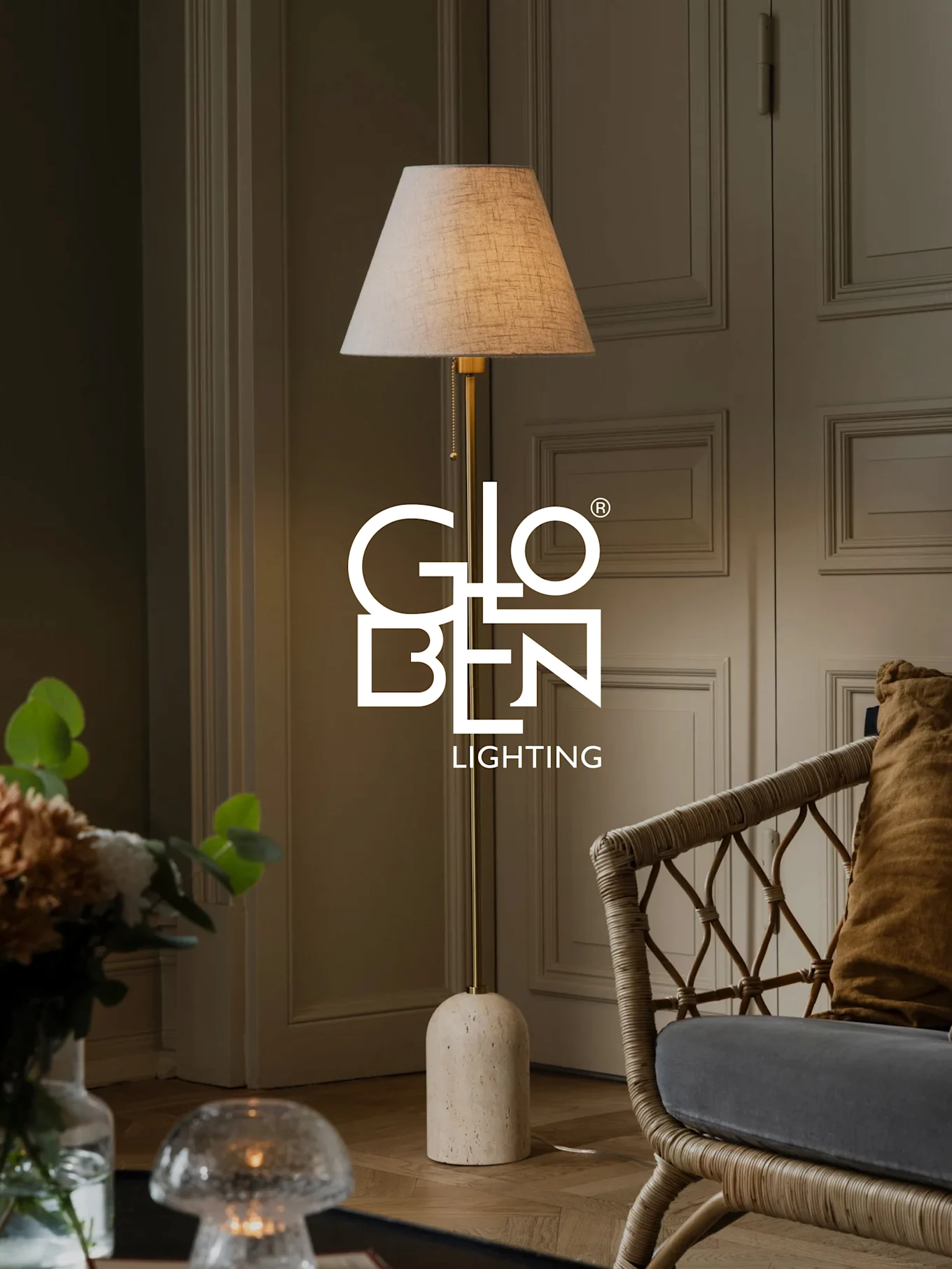 En golvlampa med beige skärm, mässingsstativ och rundad stenbas står bredvid en rottingstol.