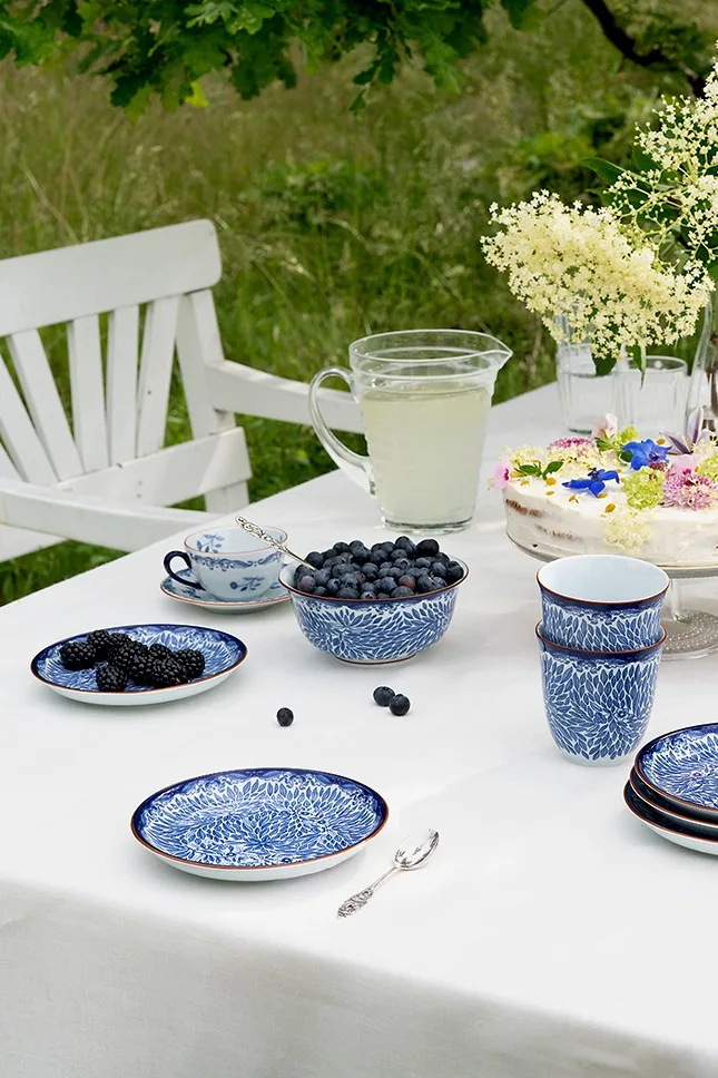Utomhus dukat bord med blåvita mönstrade rätter, färska blåbär och björnbär, en lemonadkanna och en blomsterdekorerad tårta.