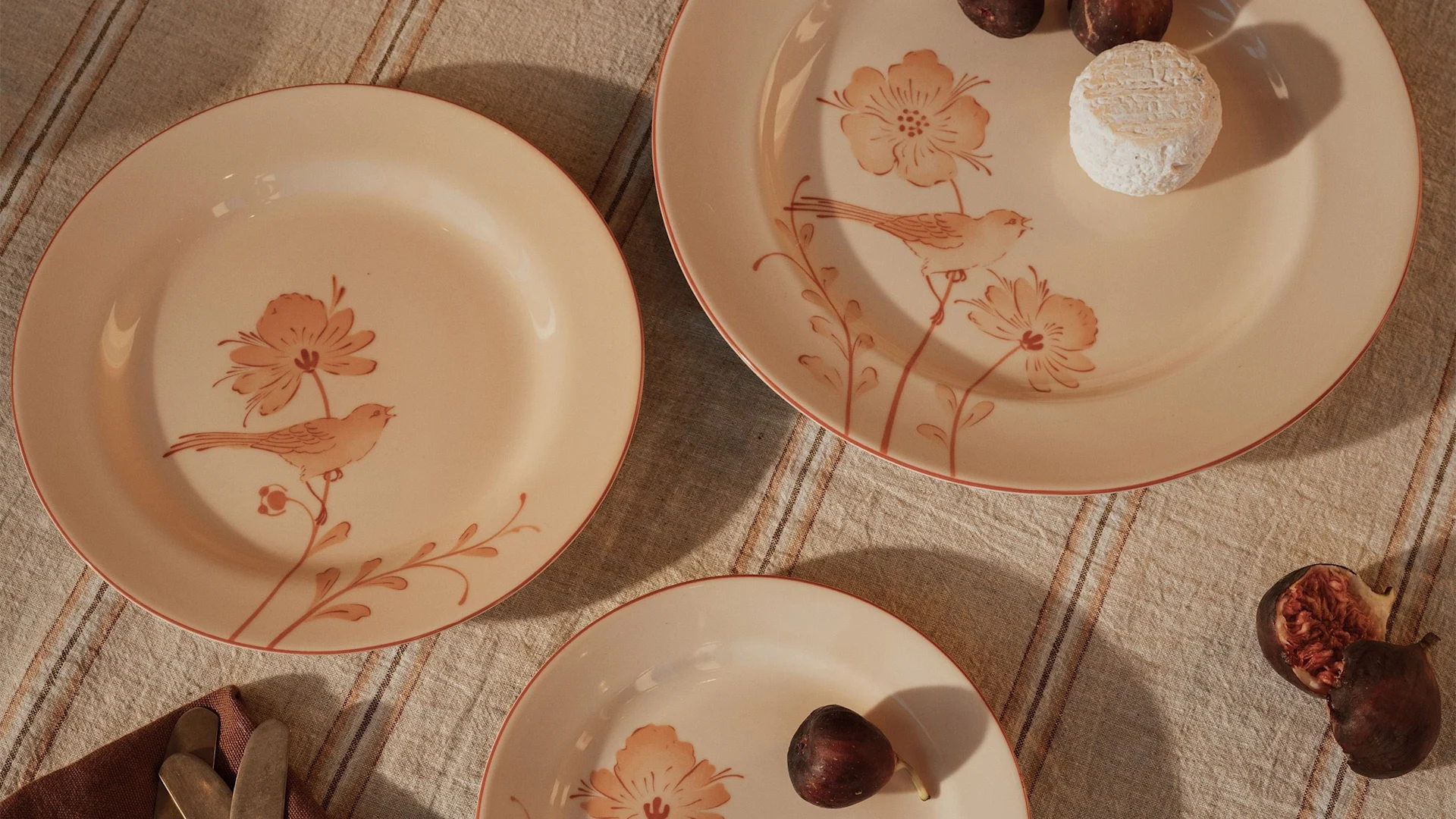 Tre ljusa persikofärgade tallrikar med fågel- och blommotiv på en randig duk, med fikon och en ost från Rörstrand. Tallrikarna kommer ifrån Jubilee fågel kollektionen.