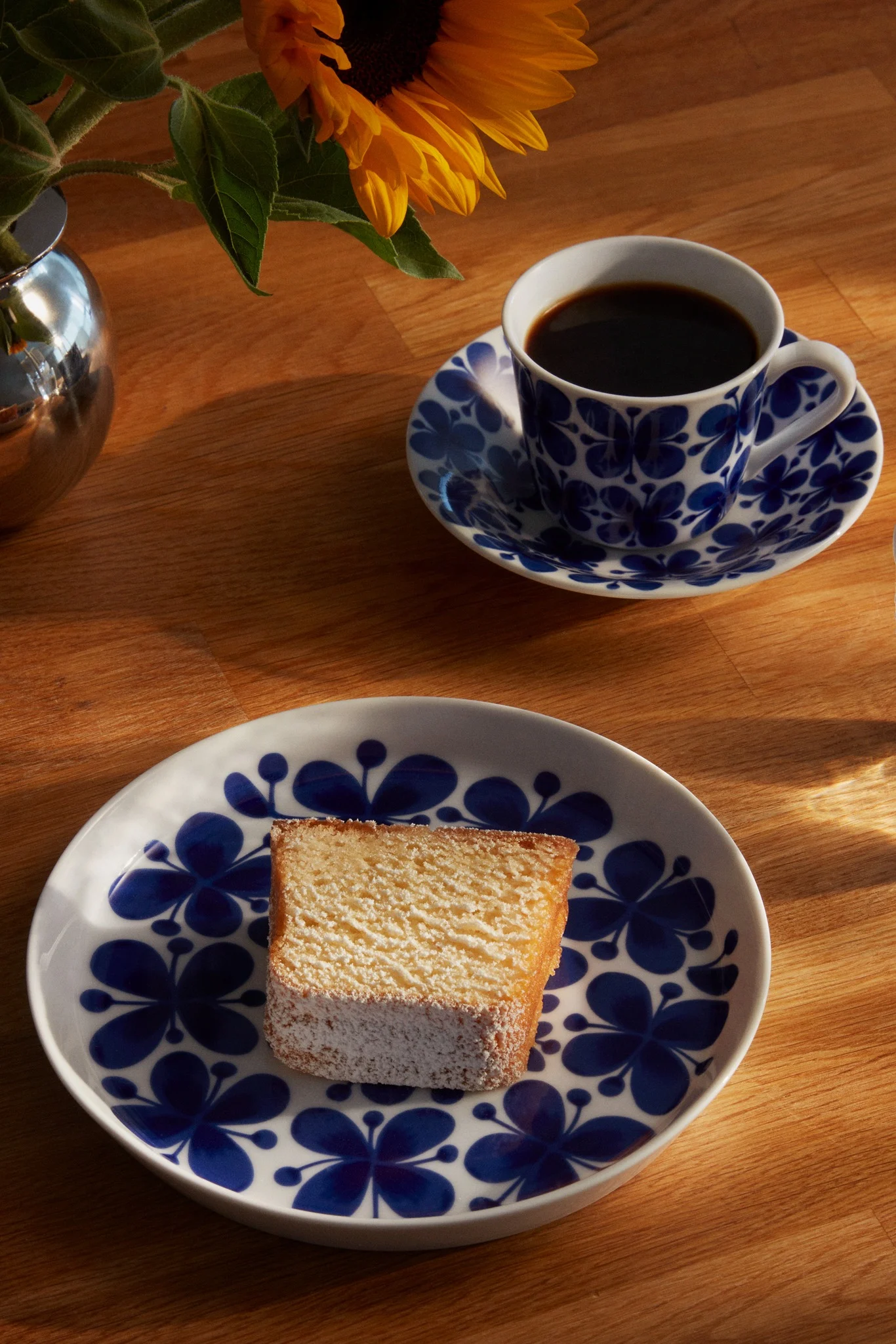 Puderkaka på en blå blommig tallrik, svart kaffe i en matchande kopp och en solros i en silvervas på trä.