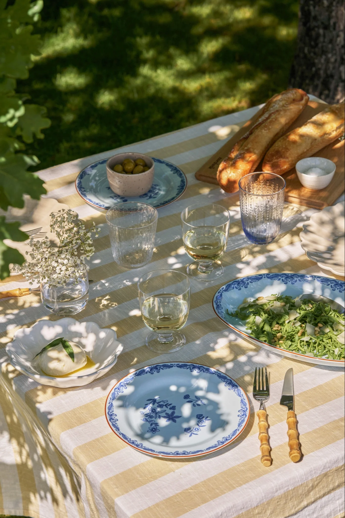 Ett utomhuspicknickbord dukat med en gul och vitrandig duk, burrata, sallad, baguetter, oliver och blåmönstrade tallrikar.