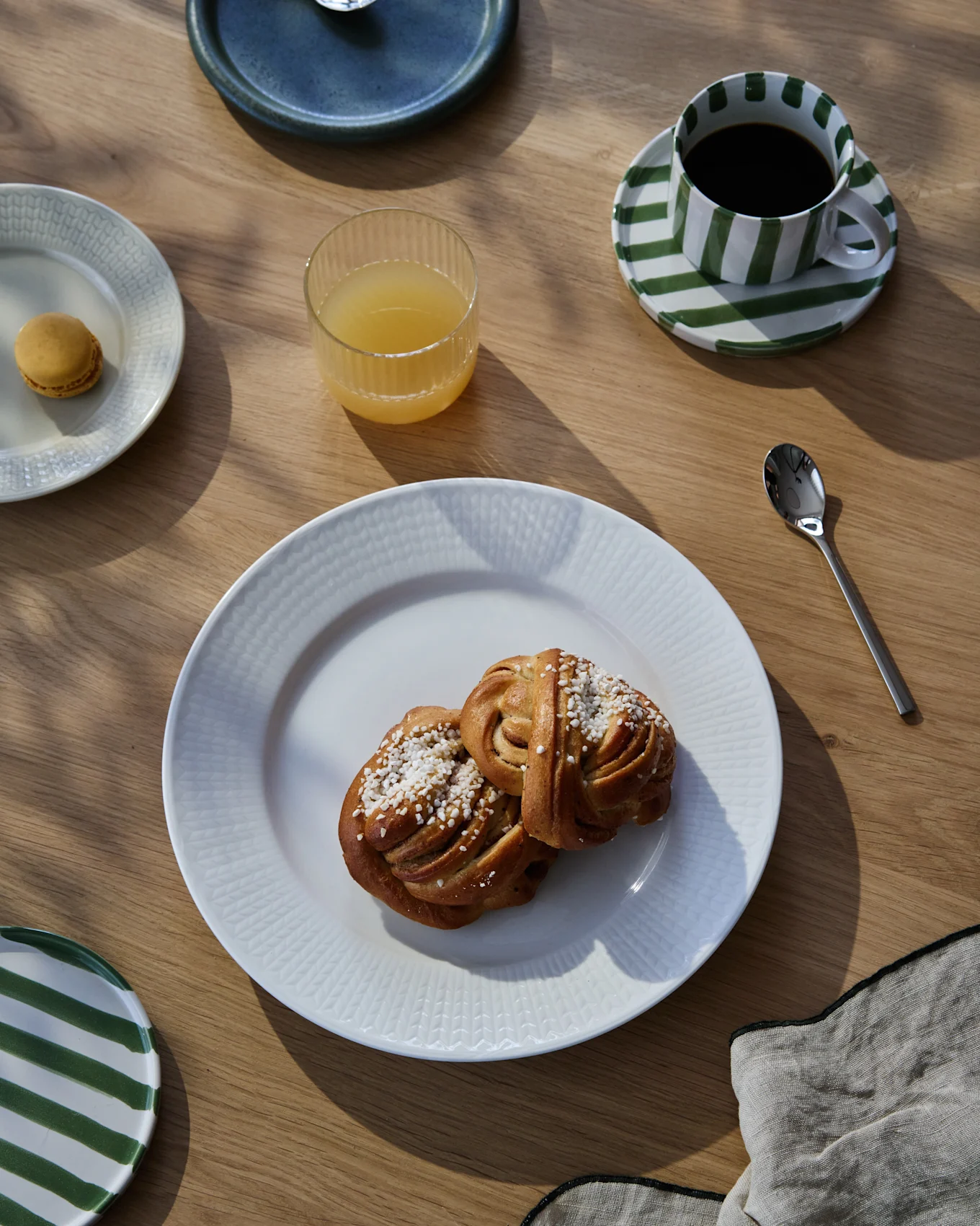 En frukostdukning på ett träbord med två kanelbullar, kaffe i en randig kopp, apelsinjuice och en makaron.