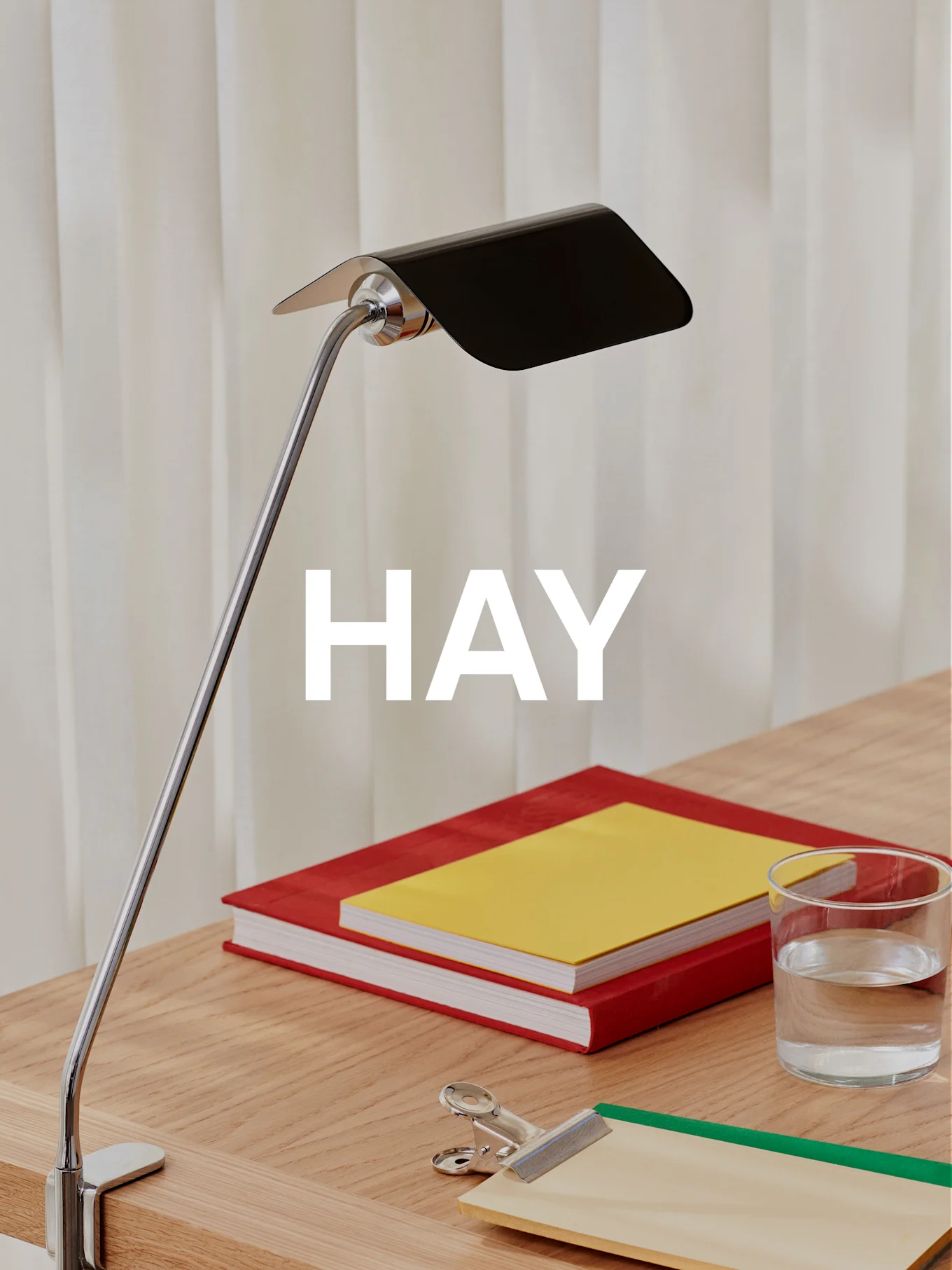 En kromad skrivbordslampa med svart skärm lyser upp ett träskrivbord med två böcker, ett glas vatten och en urklippstavla.