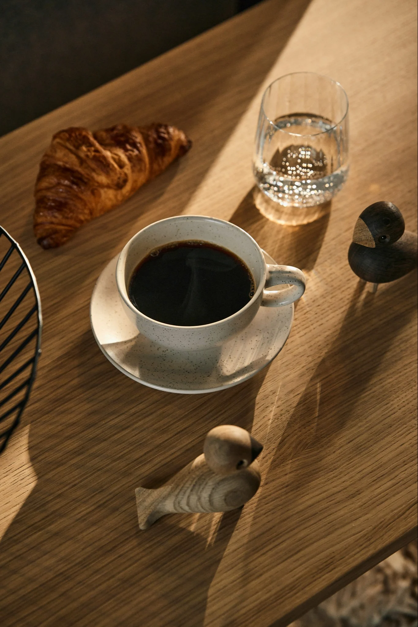 En frukostdukning med kaffe, en croissant, vatten och två träfågelfigurer på ett soligt träbord.