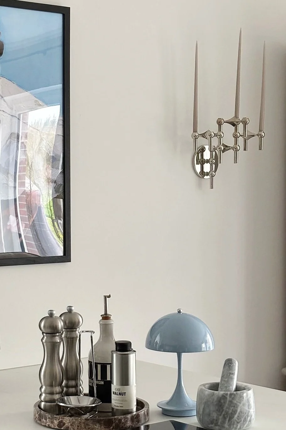 En silverfärgad vägglampa med tre ljus hänger ovanför ett vitt bord med köksredskap: kvarnar, oljeflaskor, en blå svamplampa och en marmormortel.