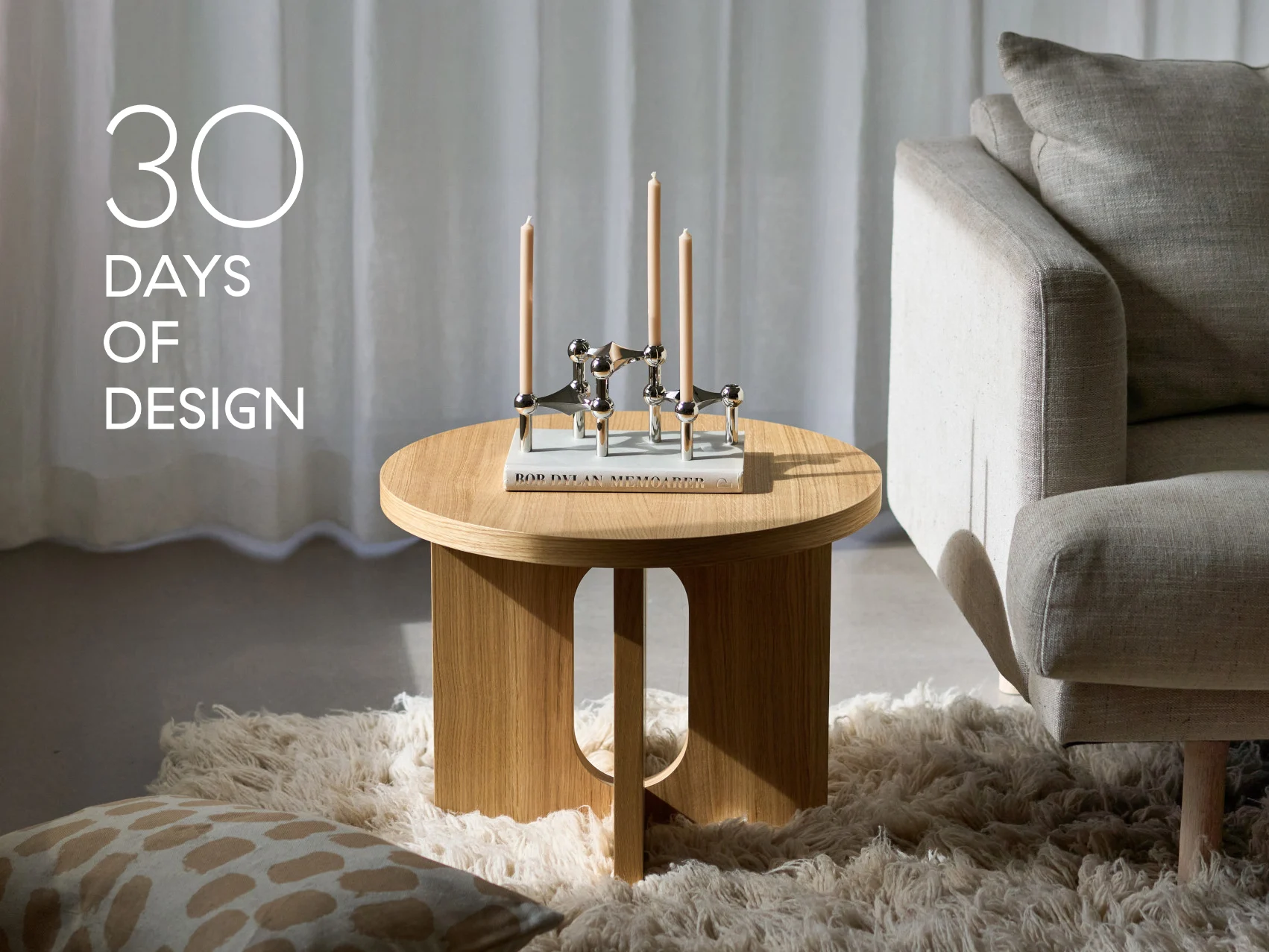 Modernt vardagsrum med runt sidobord i trä, silverljusstake med tre ljus, en bok och en beige soffa. Text: '30 DAGAR AV DESIGN'.