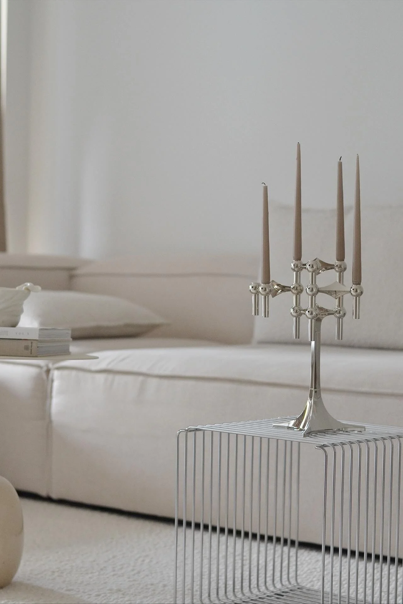 En silverfärgad kandelaber med fem ljusbruna ljus står på ett minimalistiskt sidobord i metall. En ljus soffa är i bakgrunden.