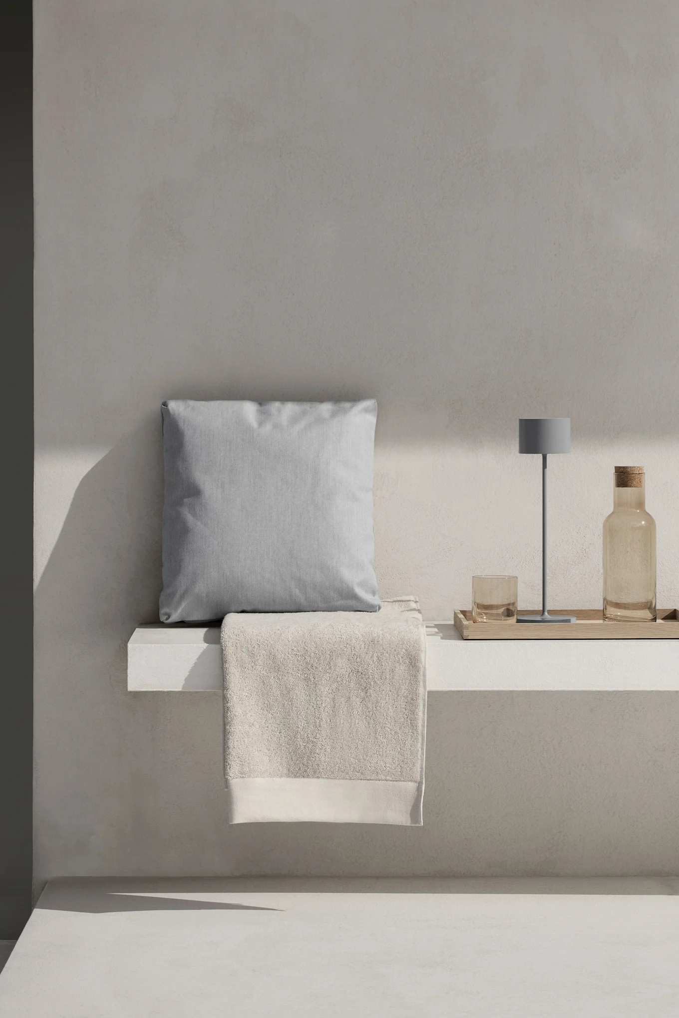 En ljusgrå kudde och beige handduk på en vit hylla, med en grå lampa, glas och flaska på en träbricka.
