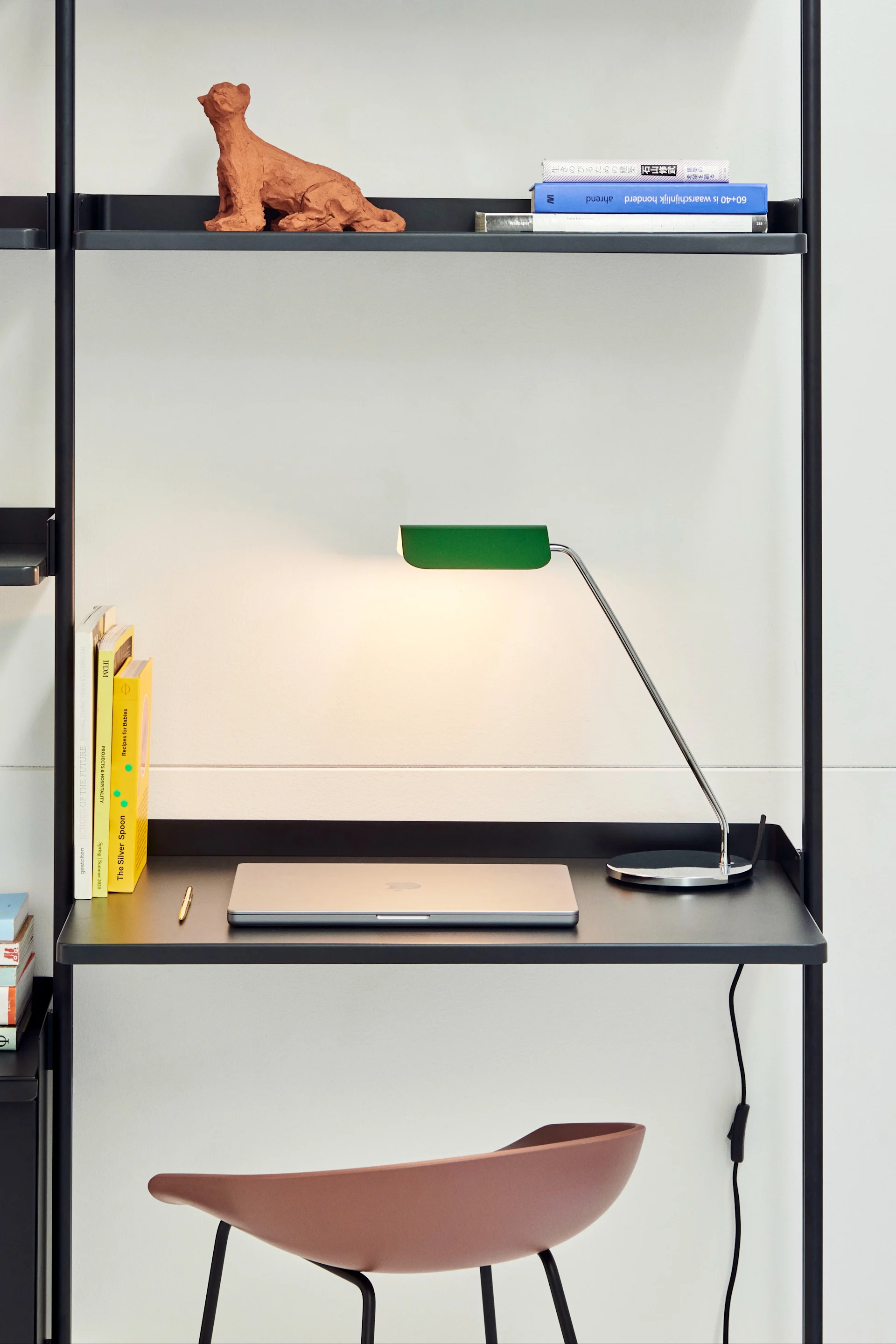 En svart modulär hyllenhet med skrivbordsdel, grön skrivbordslampa, silverfärgad laptop, böcker och terrakottaskulptur.