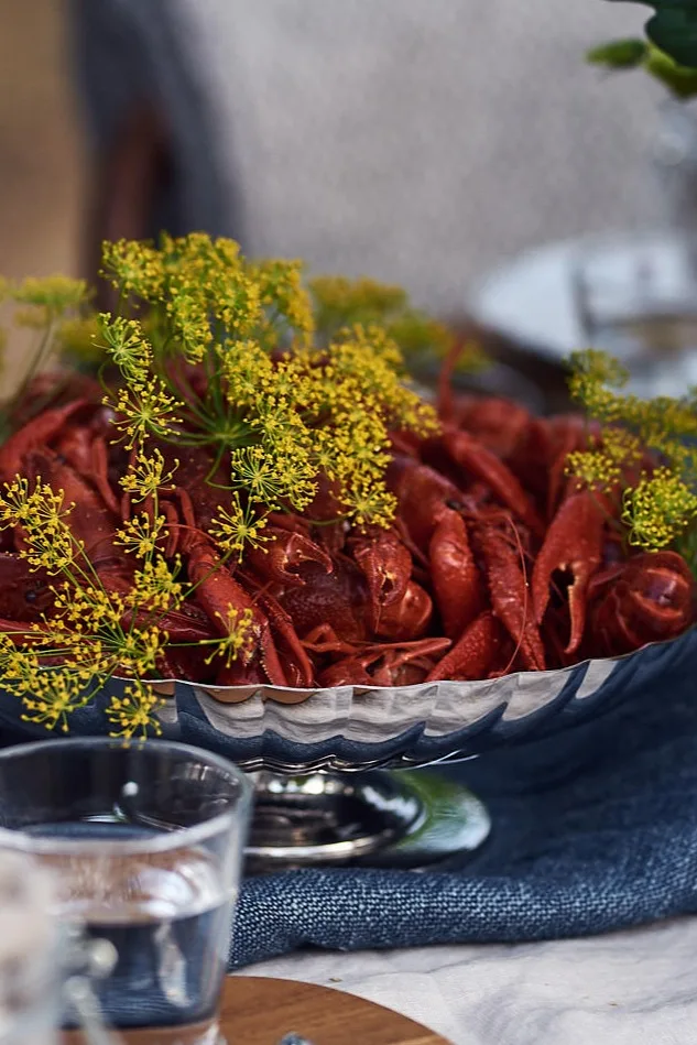 En skål med röda kräftor garnerade med gula dillblommor på ett bord med en blå duk.