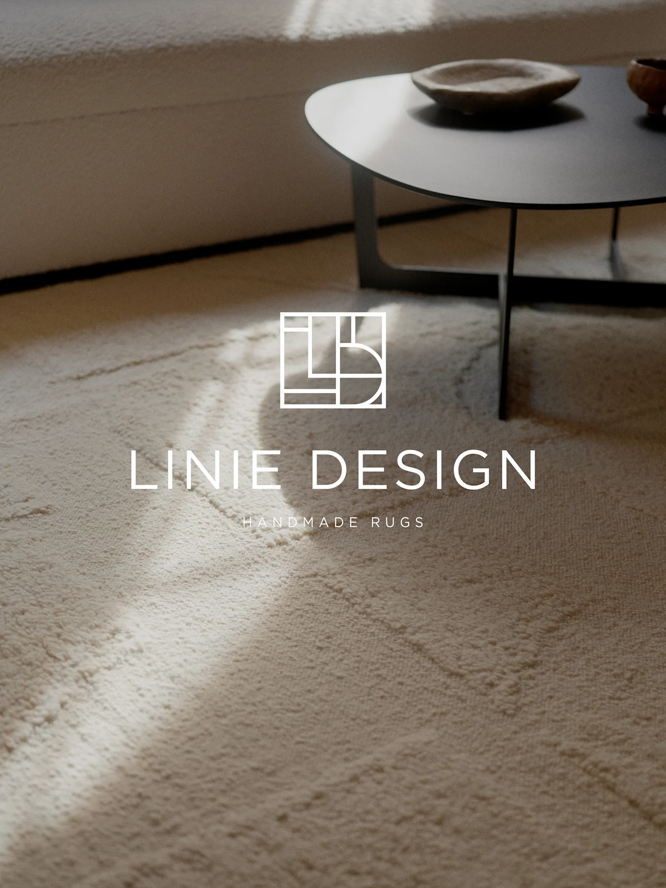 En ljus texturerad matta med Linie Design-logotypen och texten 'Handmade Rugs'. En soffa och soffbord är suddiga i bakgrunden.