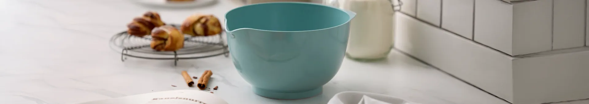 Ljusblå blandarskål, bakverk på galler, mjölkflaska och receptbok på marmorbänkskiva.