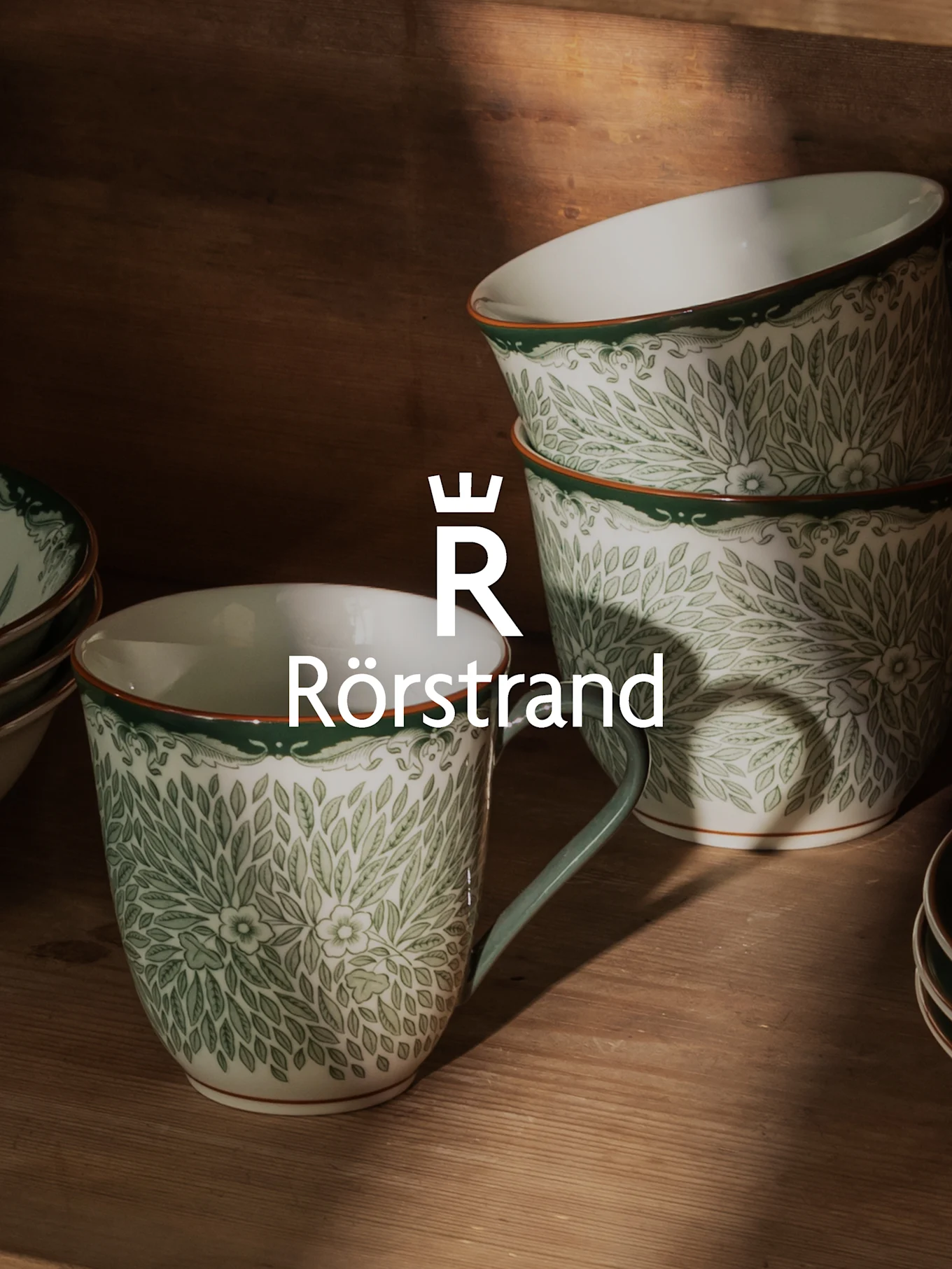 Vita keramiska muggar och skålar med grönt bladmönster, orange kant och Rörstrand-logotyp.