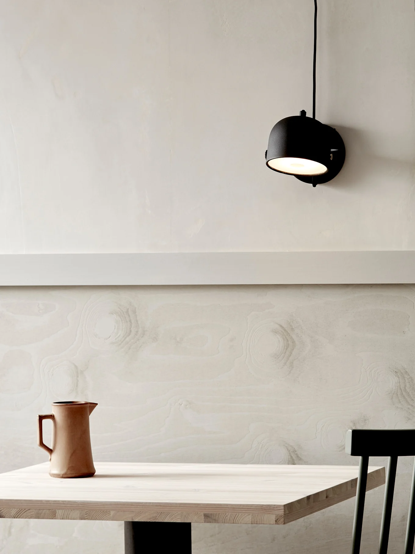 En minimalistisk interiör med ett ljust träbord, en brun kanna och en svart stol mot en texturerad ljus vägg med en svart vägglampa.