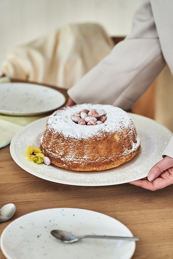 Årets påskddessert, en sockerkaka, serveras på vitt serveringsfat från Scandi Living med påskägg på och en gul blomma som dekoration. 